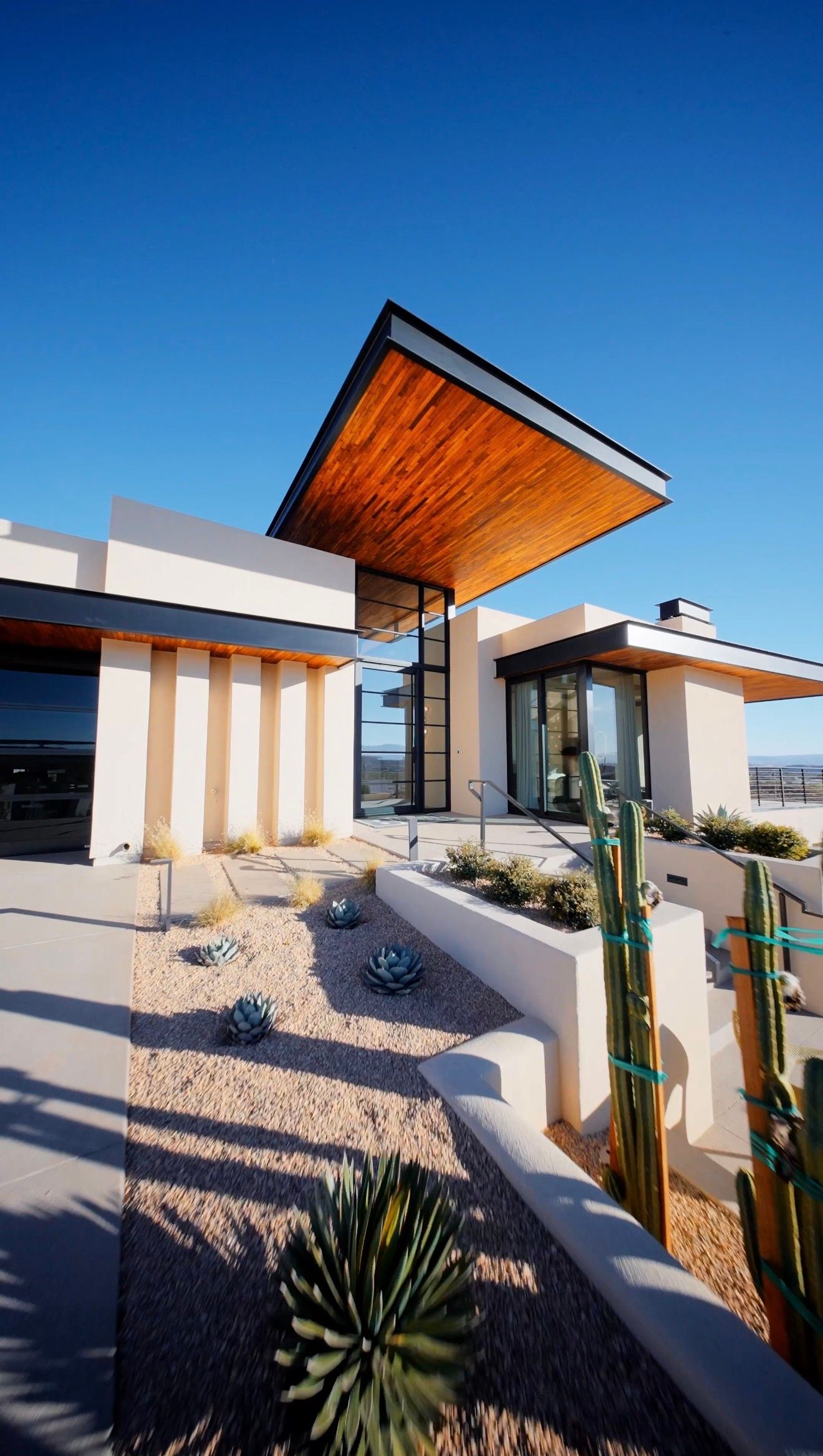 Modern desert home with wood-paneled roof overhang, white stucco walls, and drought-resistant landscaping under a clear blue sky.