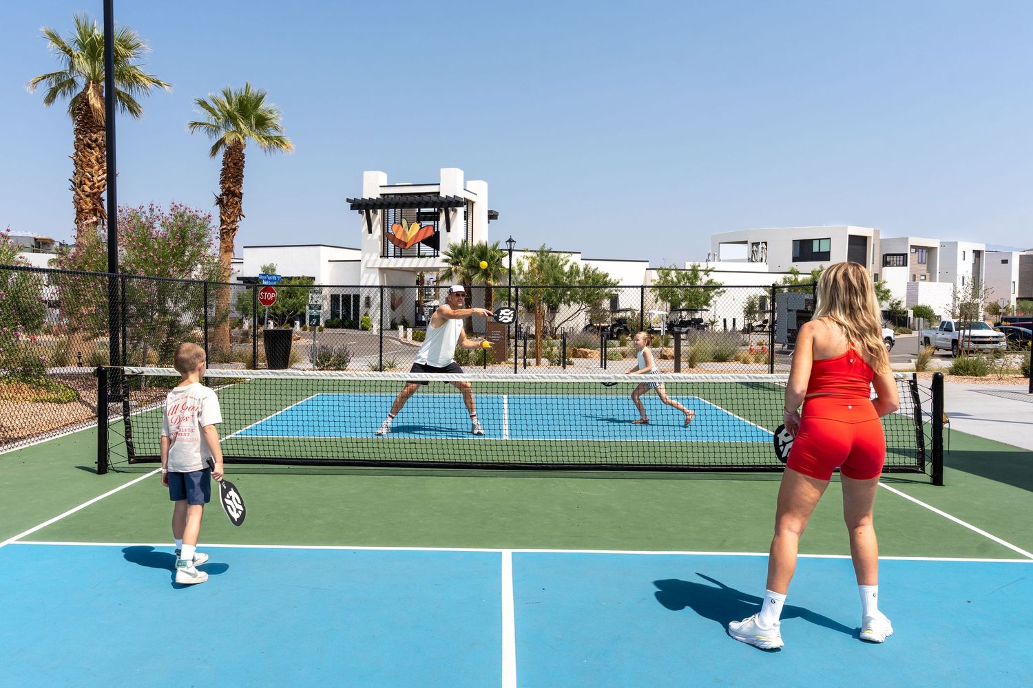 Family of four playing pickleball on a blue outdoor court at a sunny desert resort with palm trees.