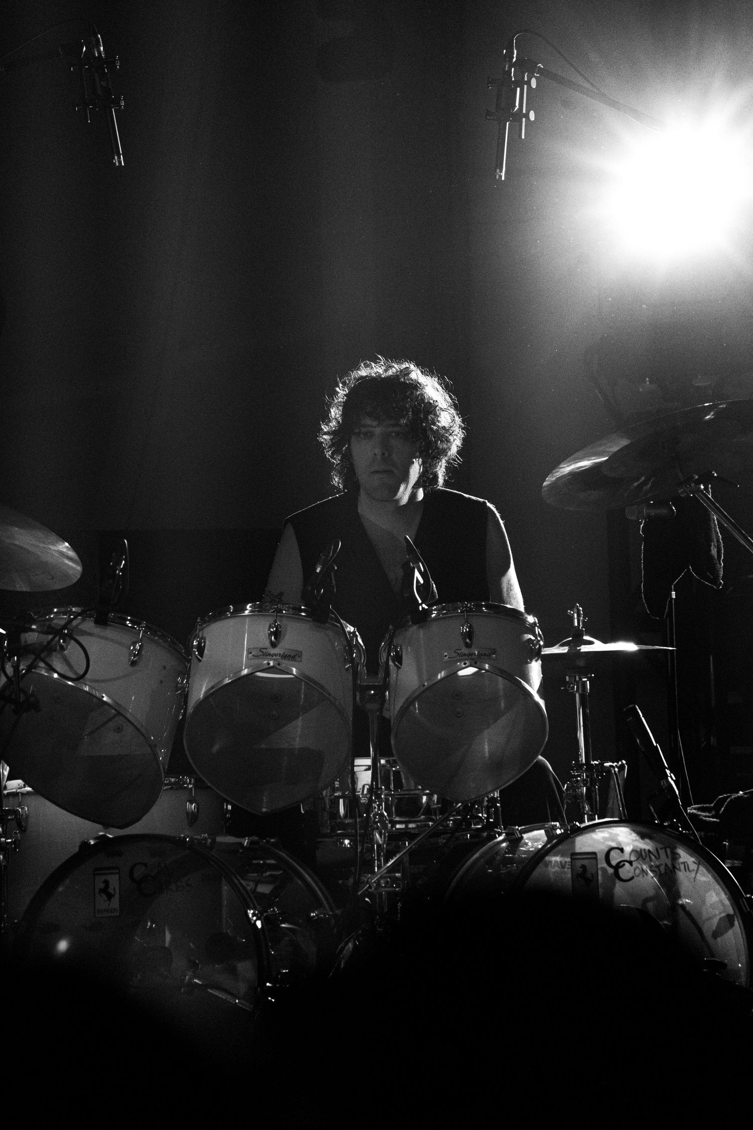 Black and white dramatic photo of a curly-haired drummer performing on stage with bright spotlight behind them.
