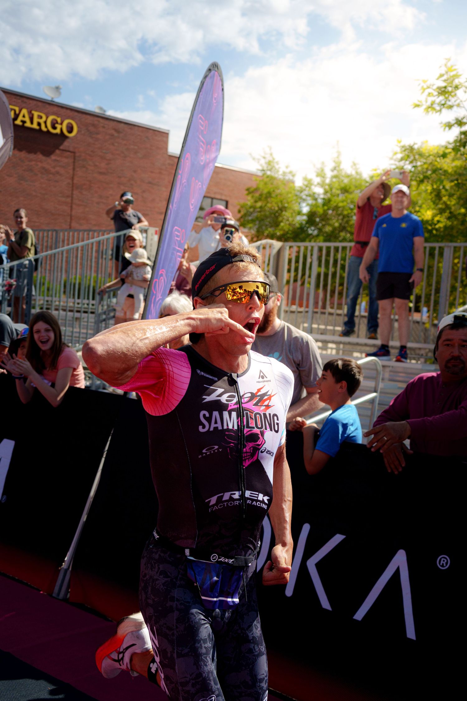 Triathlete Sam Long celebrates at the finish line of an Ironman Pro Series race, wearing sunglasses and a pink cap.