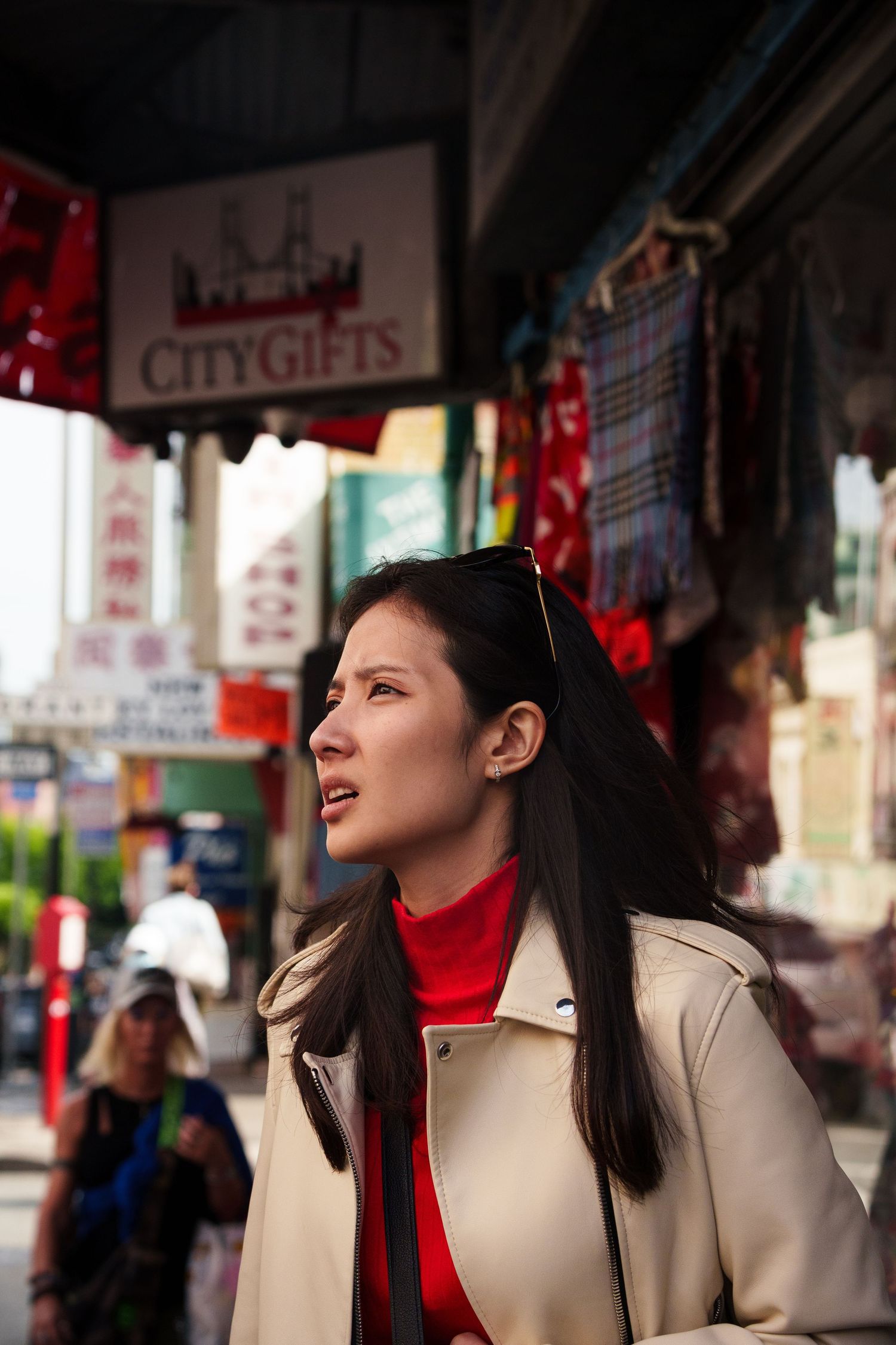 Young Asian woman in beige coat browsing a busy Chinatown street market.