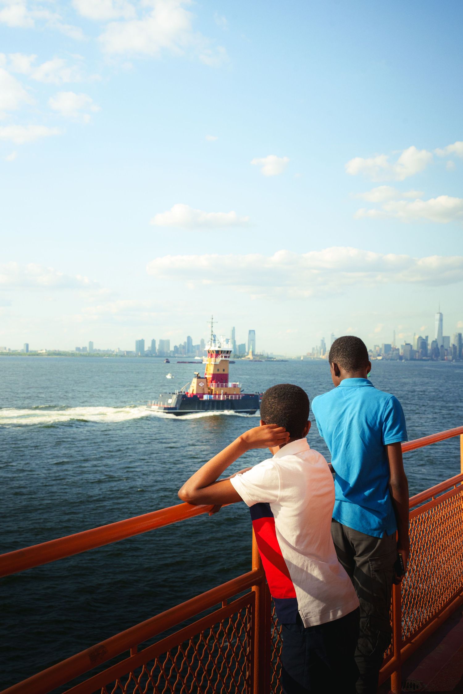 Two boys watching a tugboat from a ferry with city skyline in background.