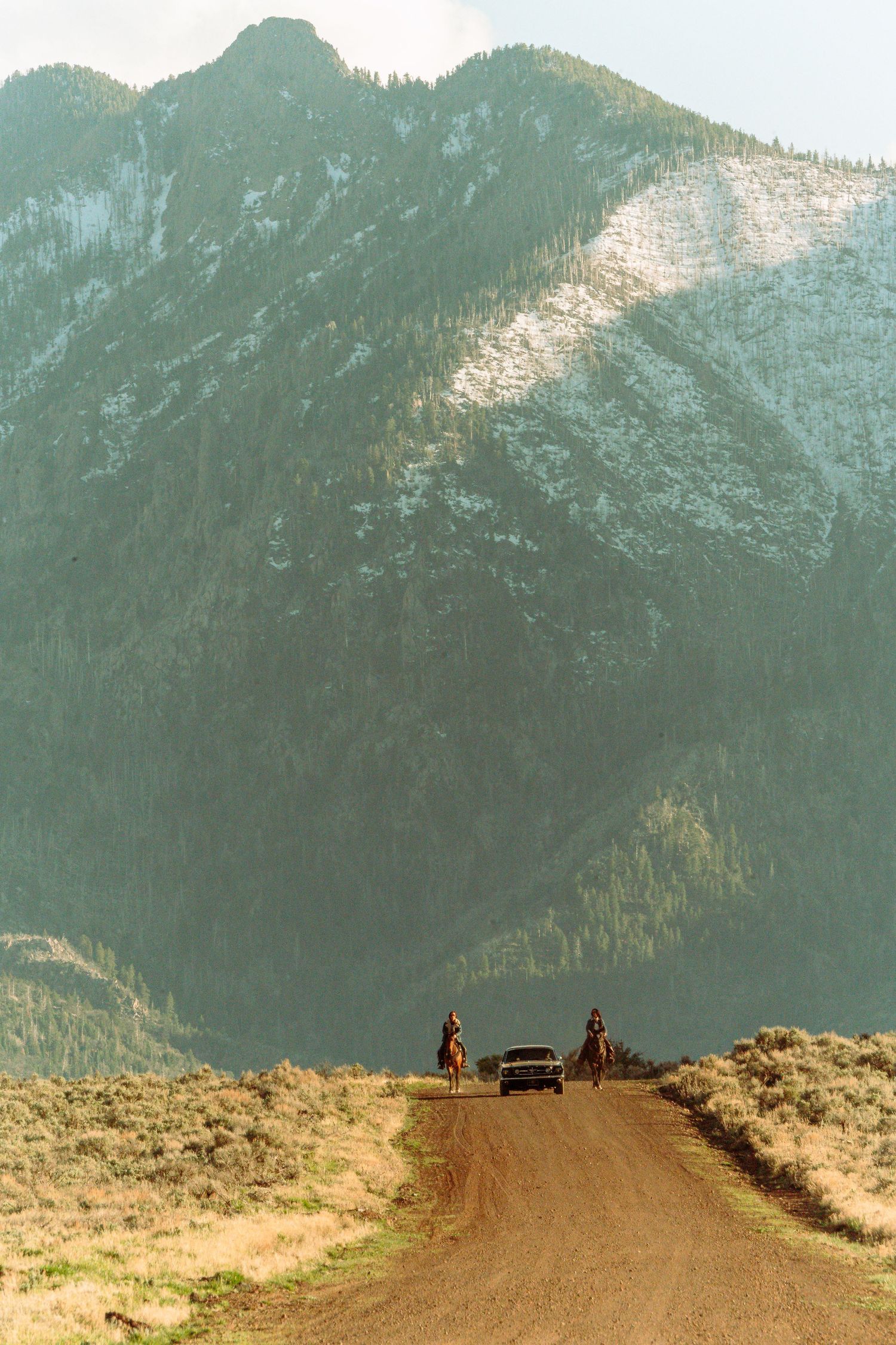 Two horseback riders flank a classic car on a dusty dirt road with a dramatic snow-capped mountain backdrop.