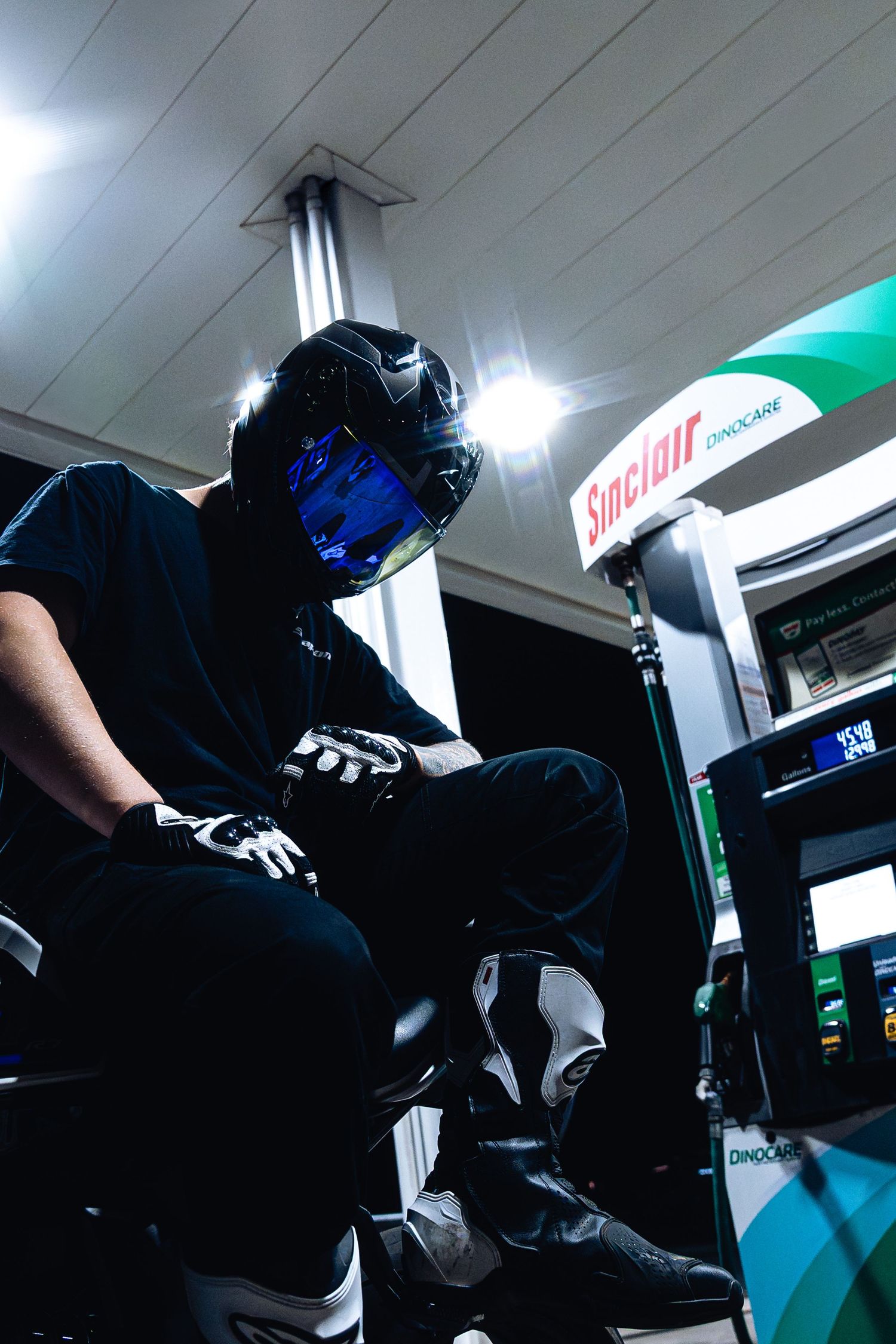 Low-angle view of helmeted motorcyclist in black gear seated on bike at Sinclair gas station.