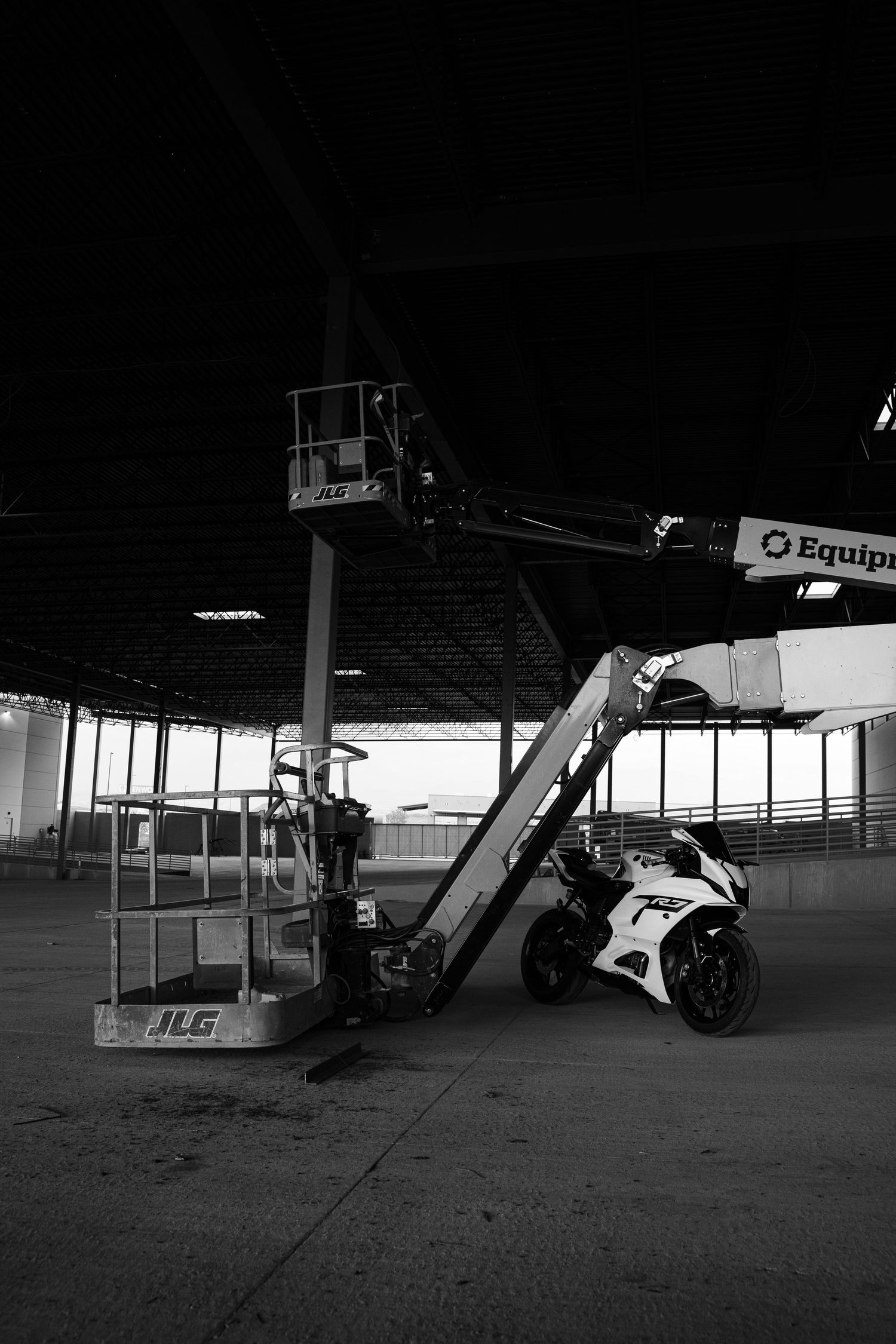 Black and white photo of a white sport motorcycle parked beside a JLG boom lift inside an empty warehouse.
