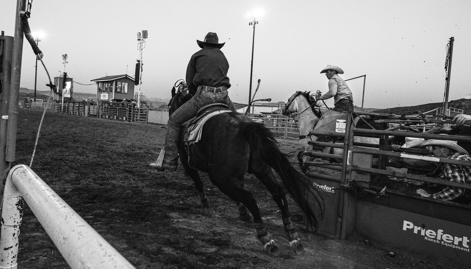 A cowboy rides a dark horse in a rodeo arena under bright lights in a black and white photograph.