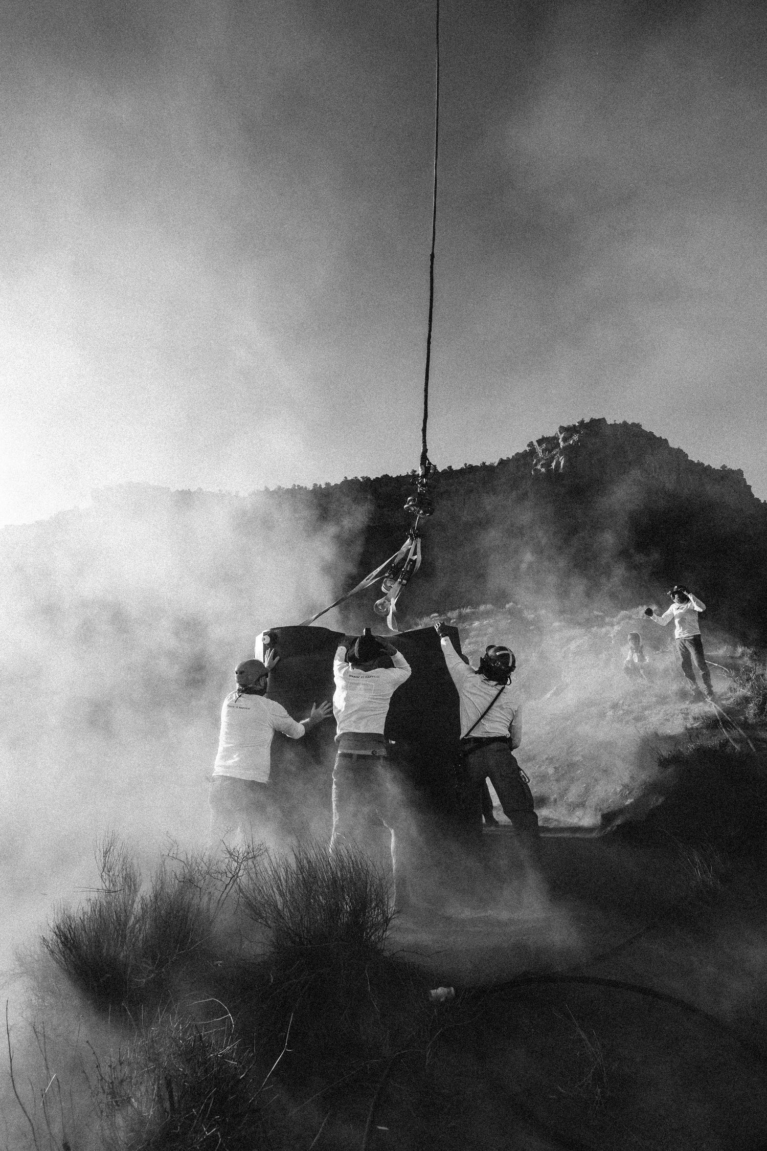 Black and white photo of crew working through smoke and fog on rocky hillside with rope.