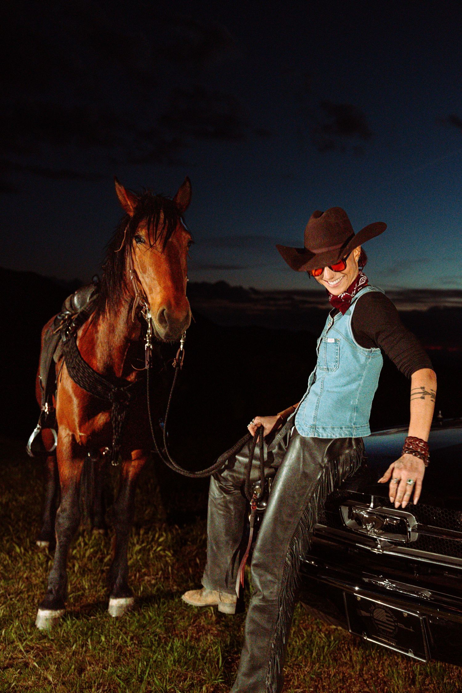 Female cowboy in hat and red sunglasses stands with saddled horse beside a classic car at night.