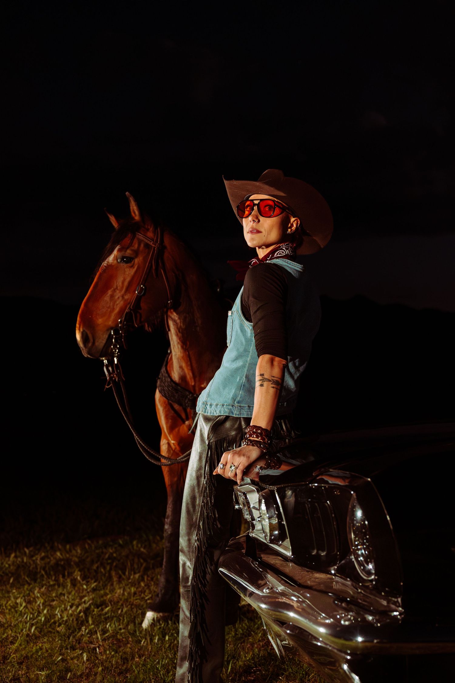 Person in cowboy hat and red sunglasses leaning on vintage car beside brown horse at night.
