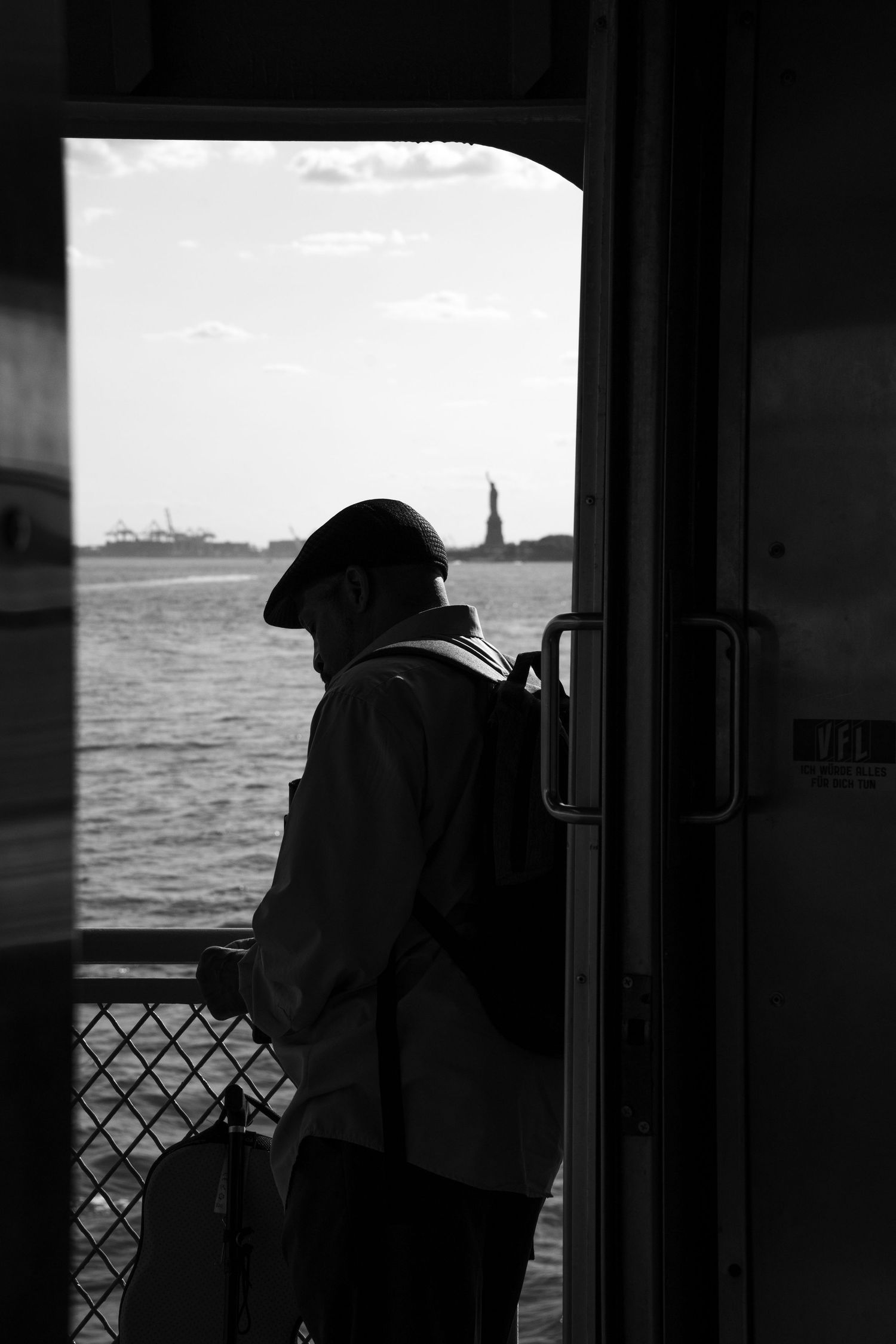 Silhouette of a man standing near a ferry door with the Statue of Liberty visible across the water in black and white.