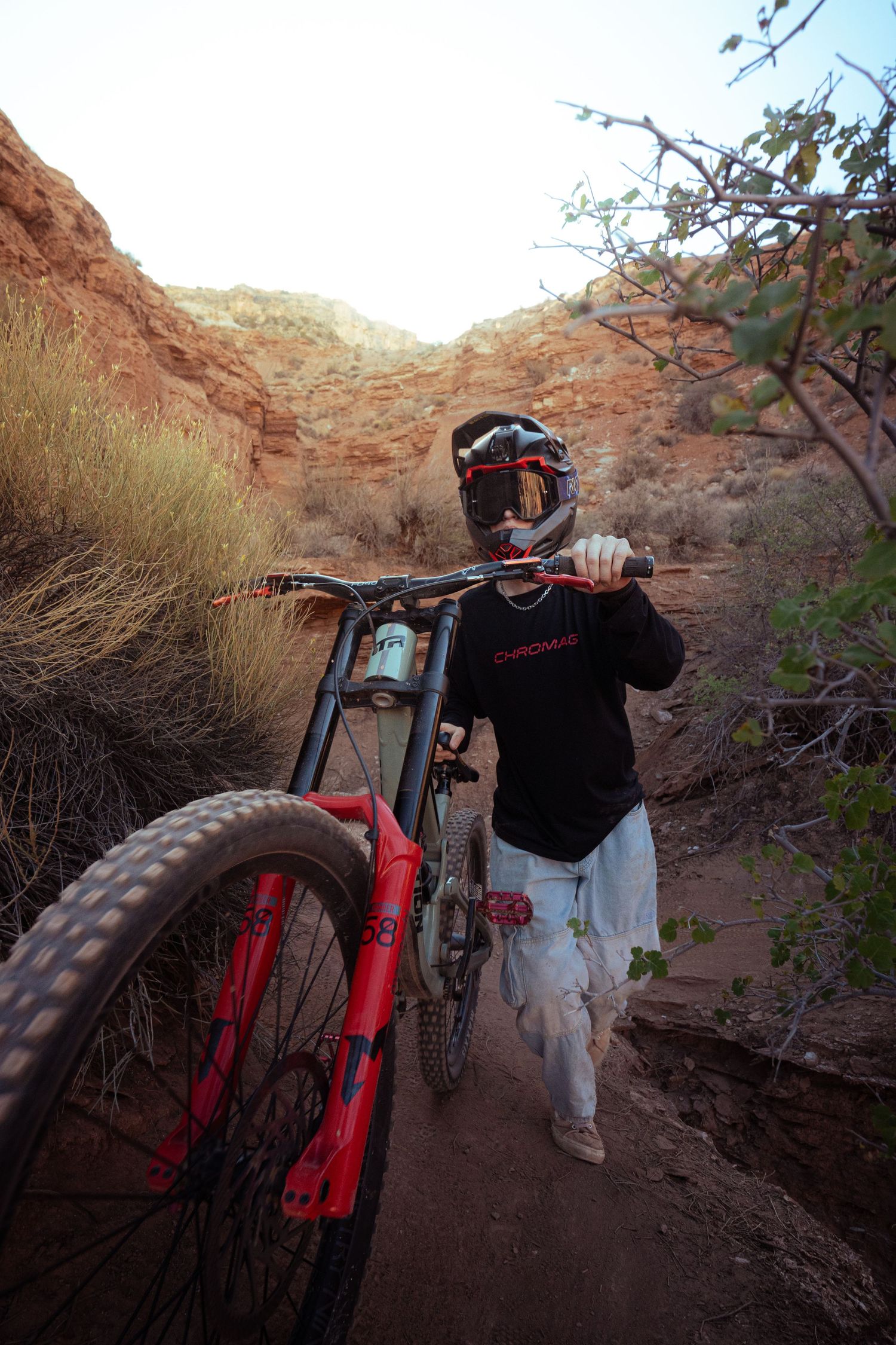 Mountain biker in helmet and goggles carries red Chromag bike through narrow desert canyon trail.