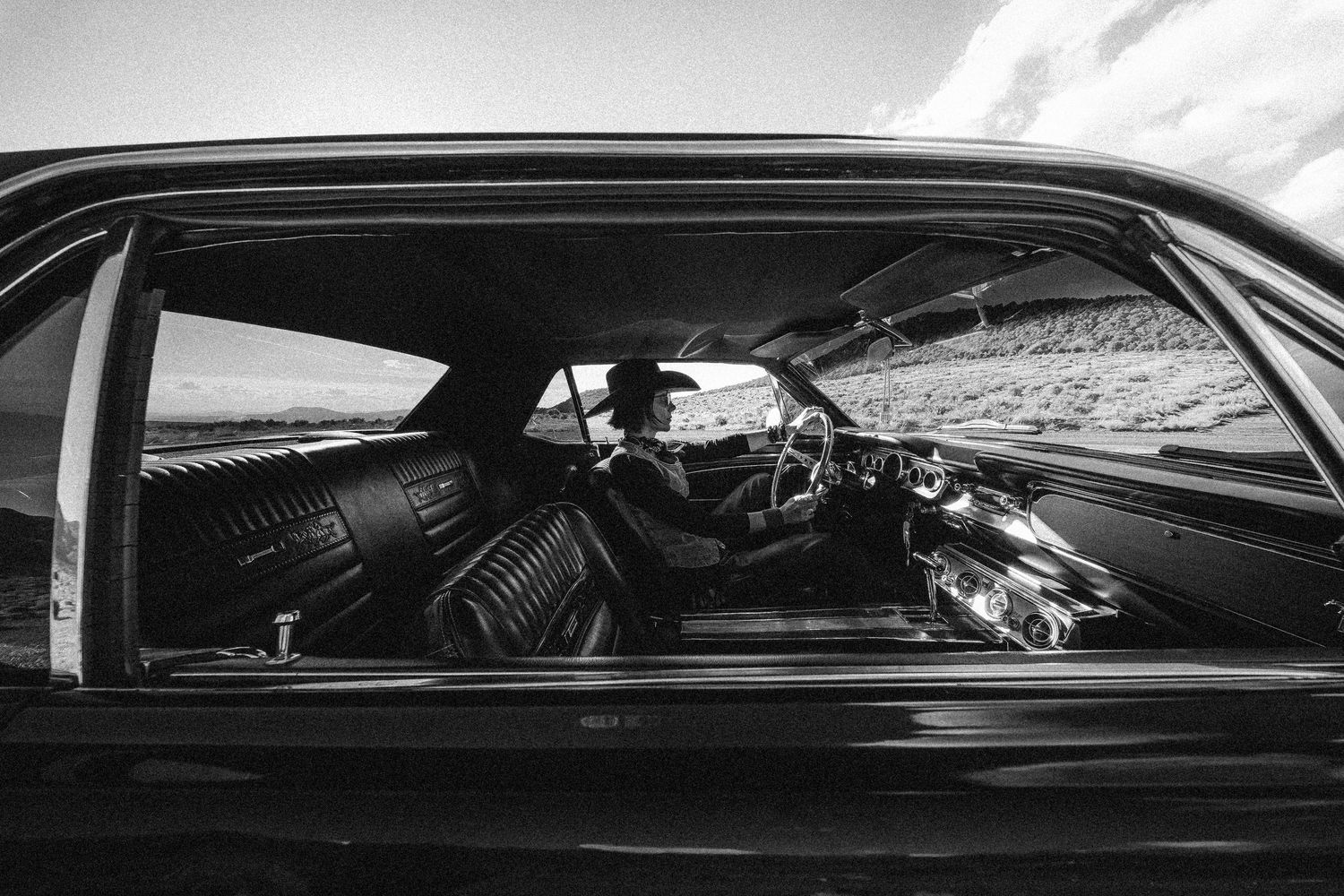 Black and white photo of a person sitting inside a classic vintage car in a desert landscape.