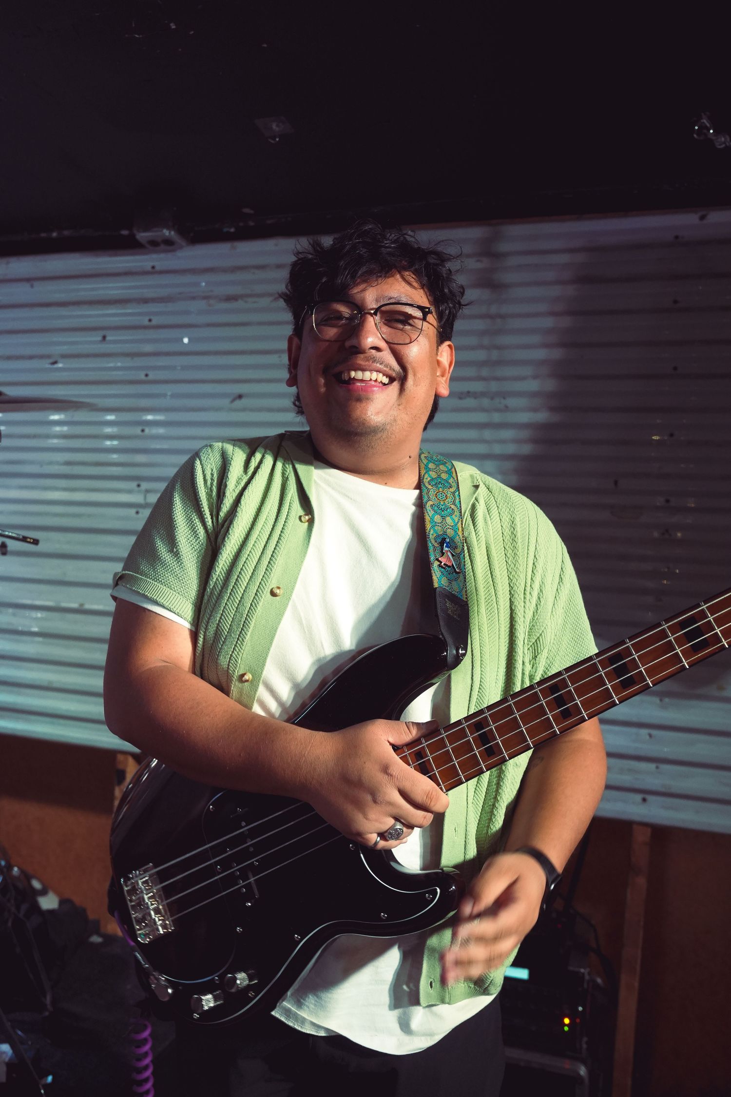 Smiling musician playing black bass guitar with decorative strap, wearing glasses and green shirt on stage.