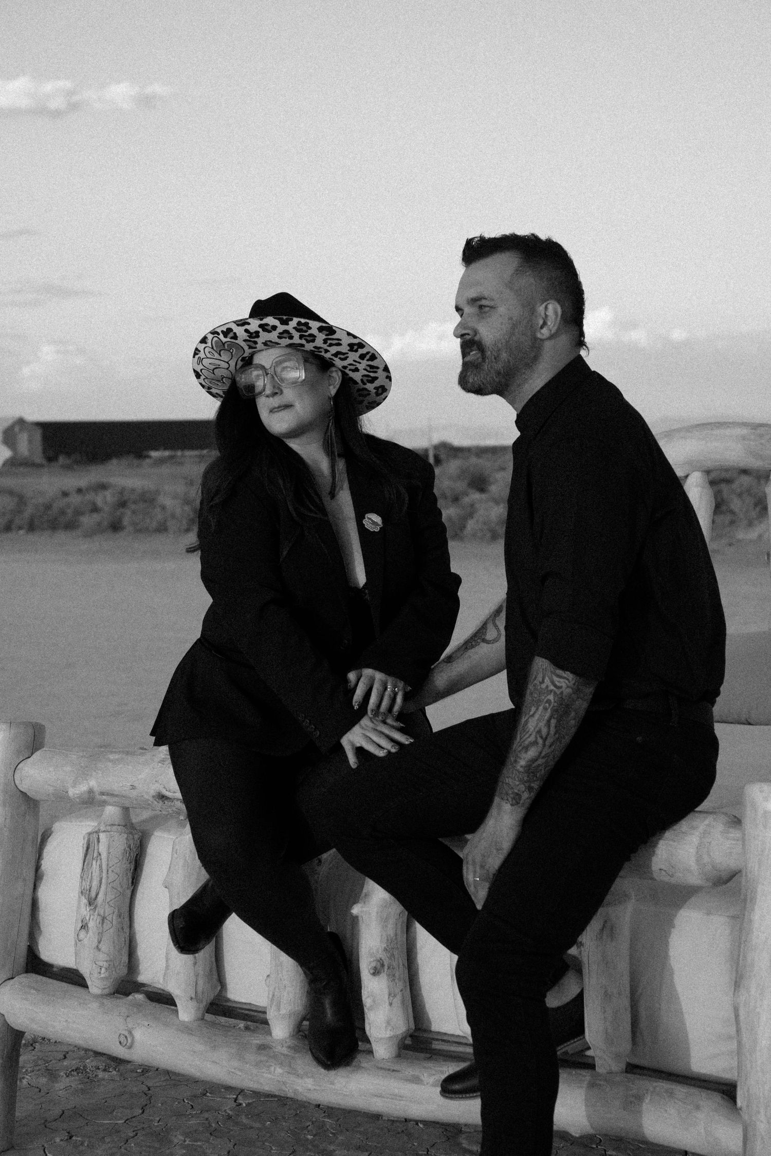 Black and white photo of couple in all black outfits sitting on bench in desert.