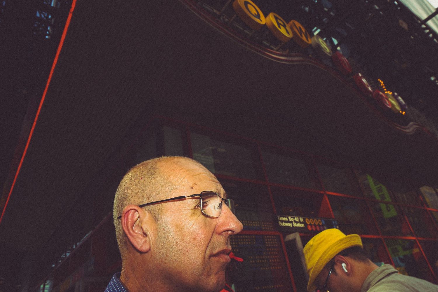 Bald man with glasses looking up at colorful neon lights in a dark urban entertainment venue.