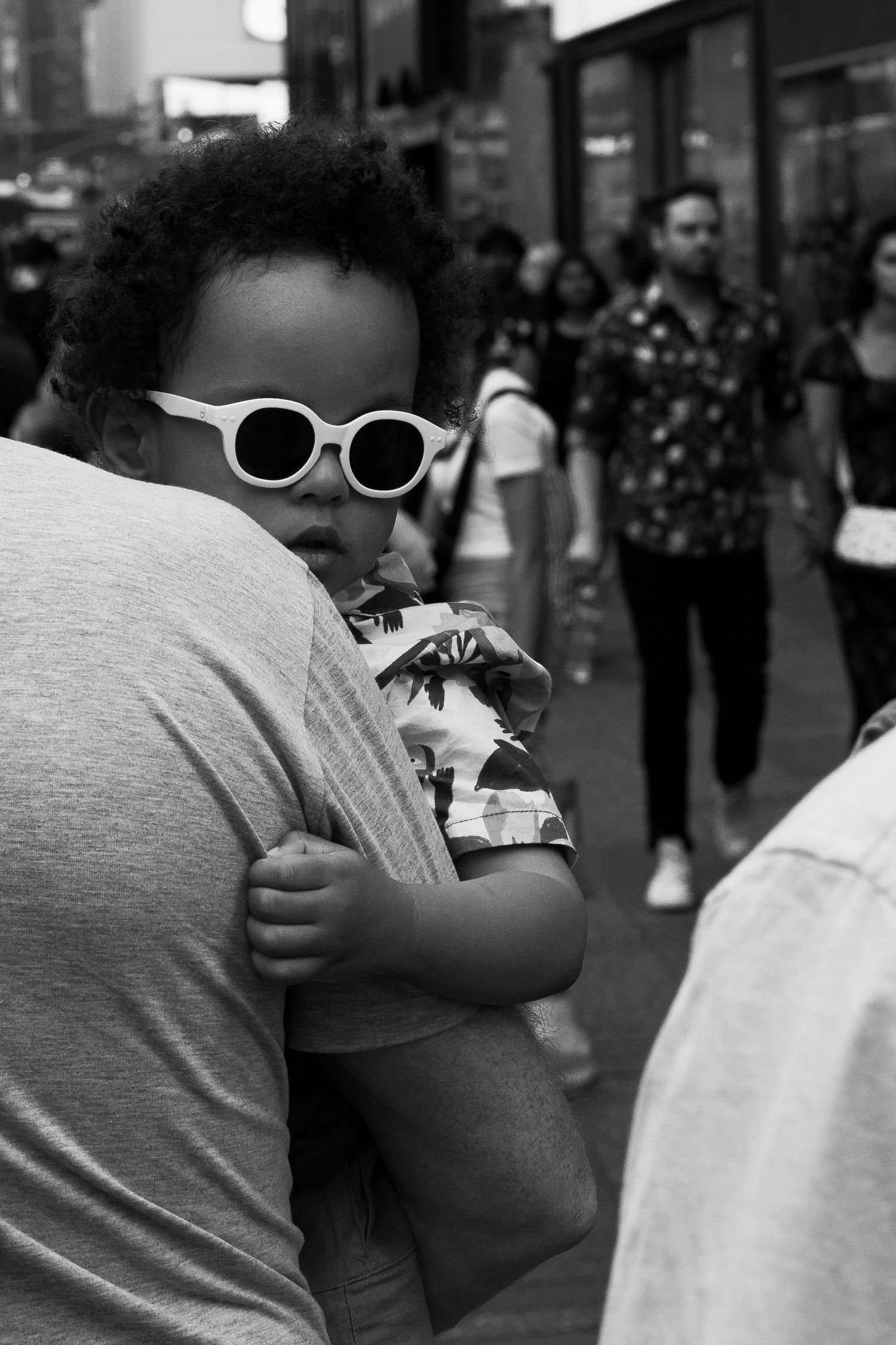 A stylish baby wearing white round sunglasses is held over an adult's shoulder on a busy city street.