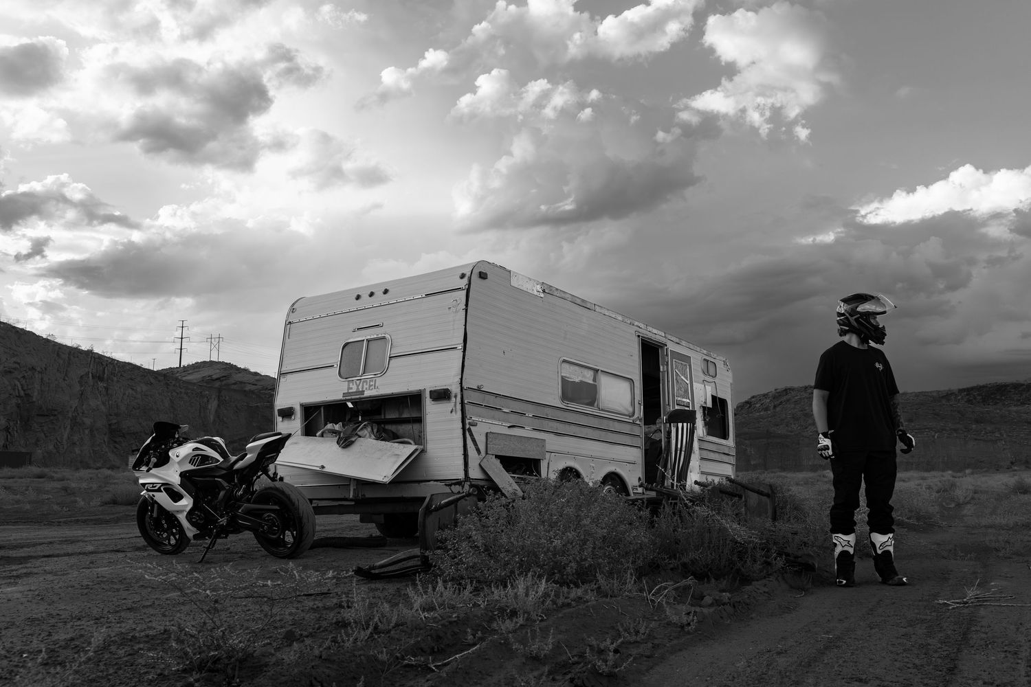 Black and white photo of motorcyclist standing beside a vintage travel trailer with a sport bike parked nearby under dramatic cloudy sky.