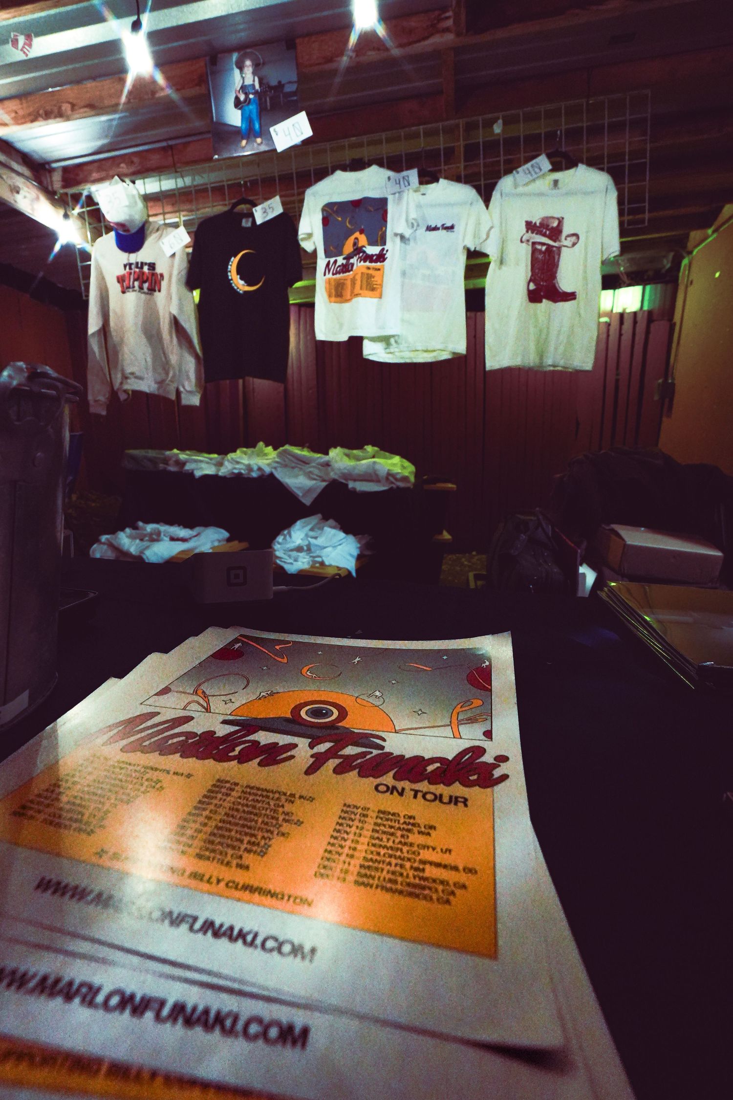 Marlon Funaki on tour merchandise table with t-shirts and tour flyers displayed in dimly lit venue.