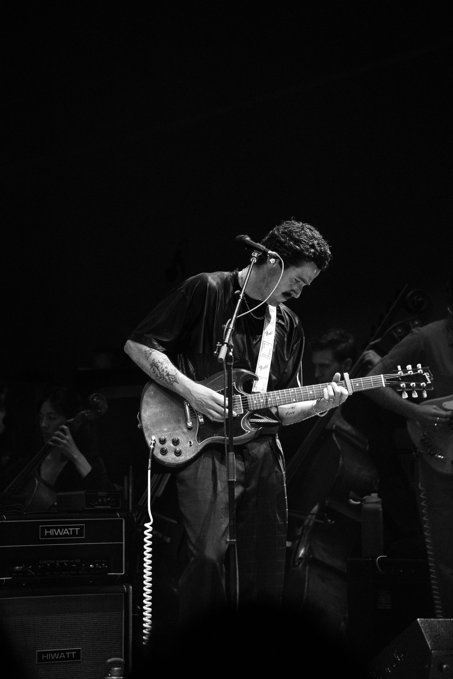 Black and white photo of a curly-haired guitarist performing on stage with Hiwatt amplifiers behind him.