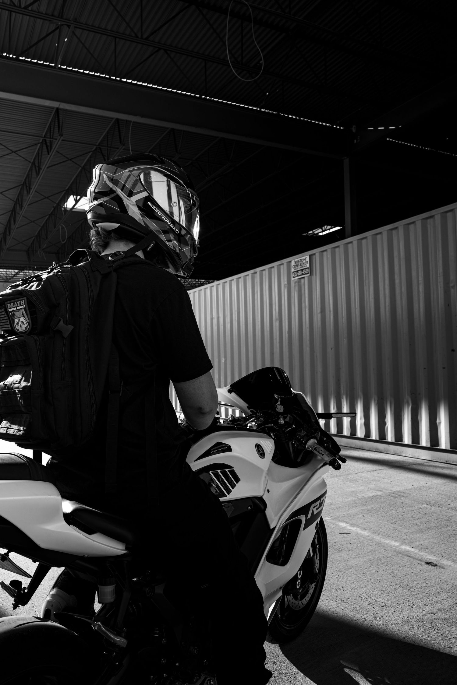 Black and white photo of motorcyclist in helmet sitting on white sport bike near corrugated metal fence.