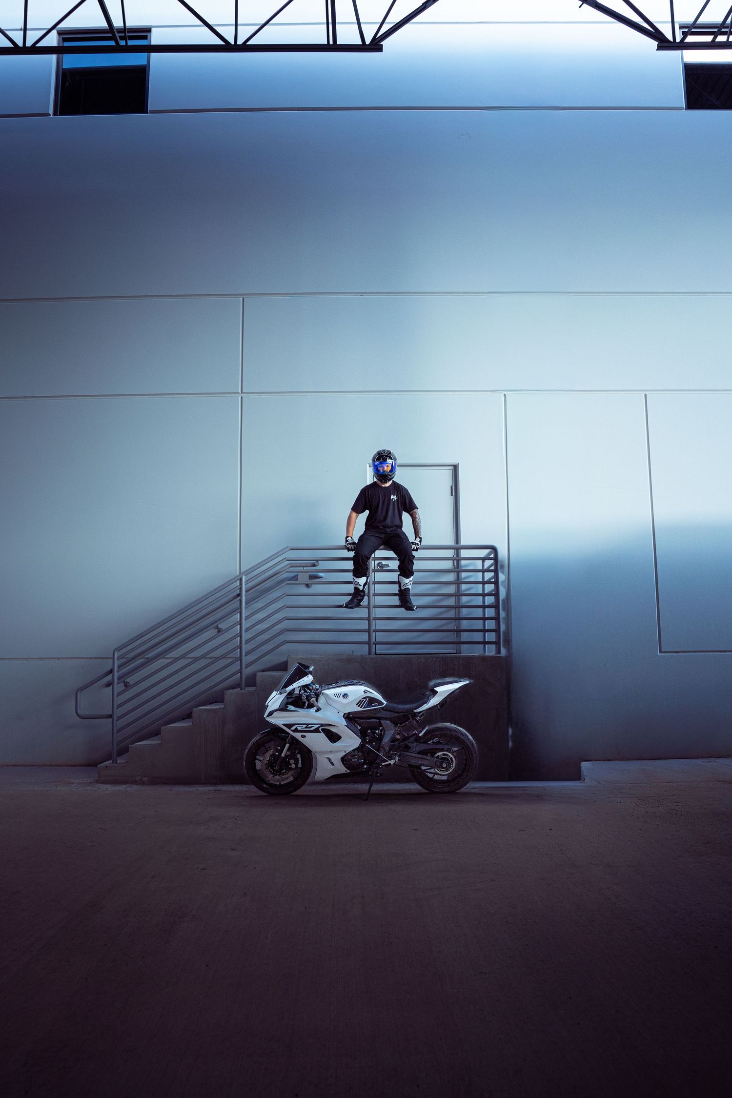 Helmeted rider jumping from staircase above white Yamaha R7 sport motorcycle in blue-lit warehouse.
