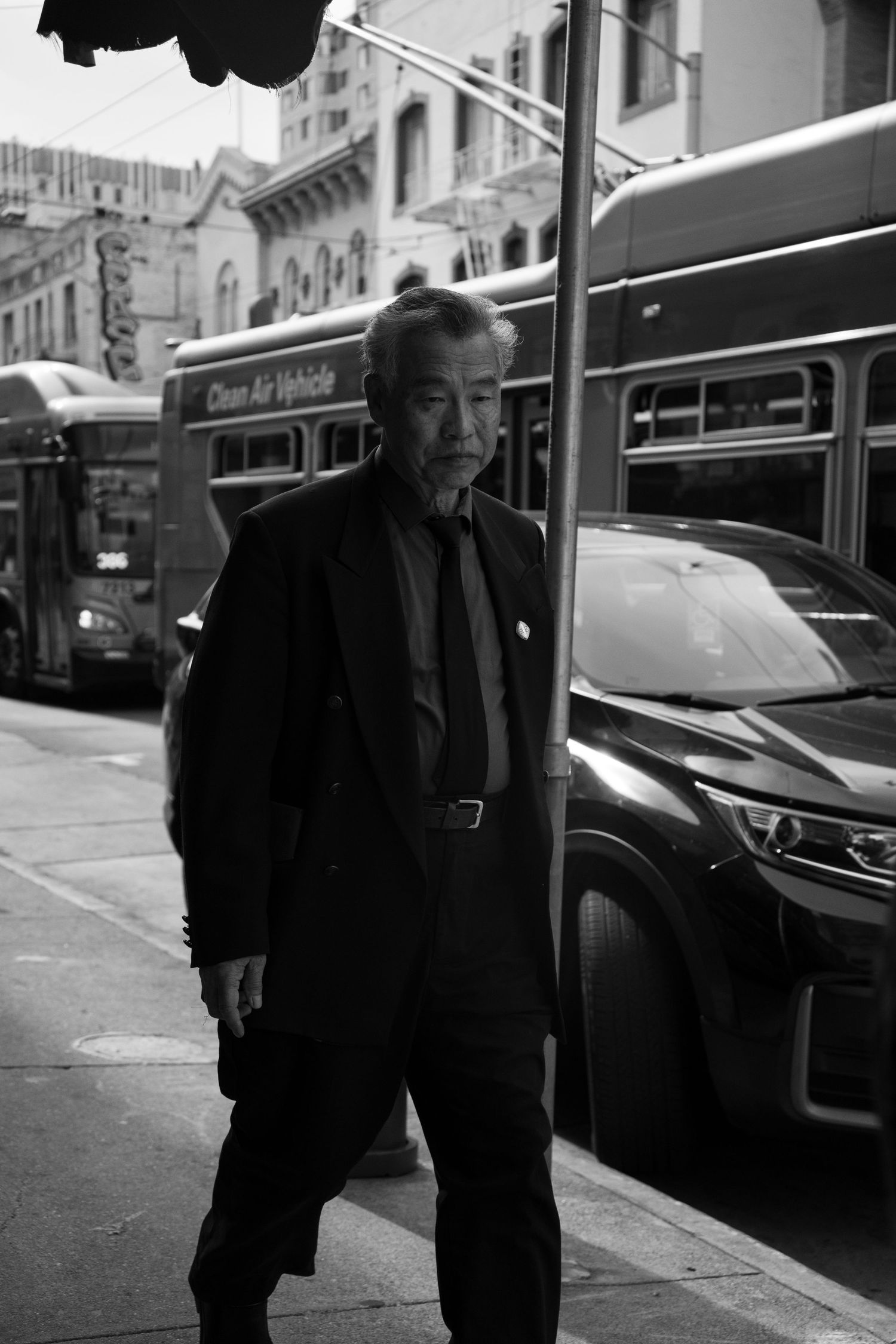 Black and white street photo of an older Asian man in a suit walking past buses in San Francisco.