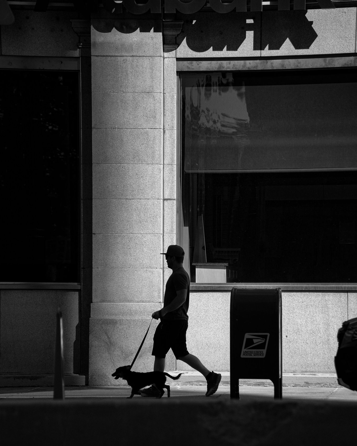 Black and white silhouette of person walking a dachshund past a storefront.