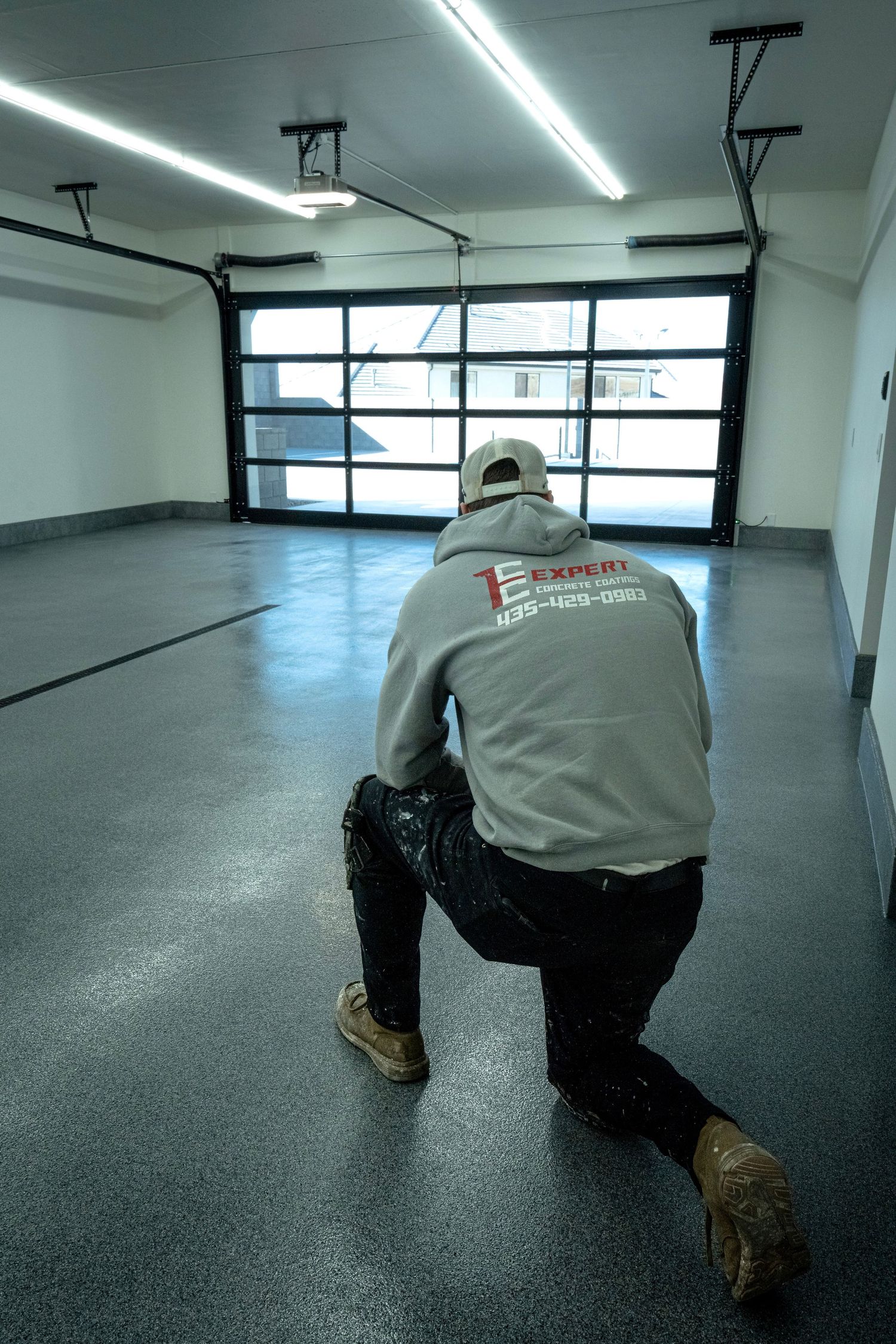 Epoxy flooring contractor kneeling on a freshly coated gray garage floor near a glass panel garage door.