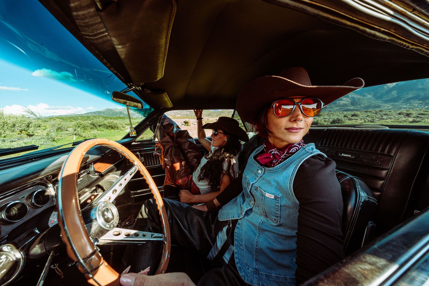A woman in a cowboy hat and sunglasses drives a classic Mustang through a scenic mountain valley.