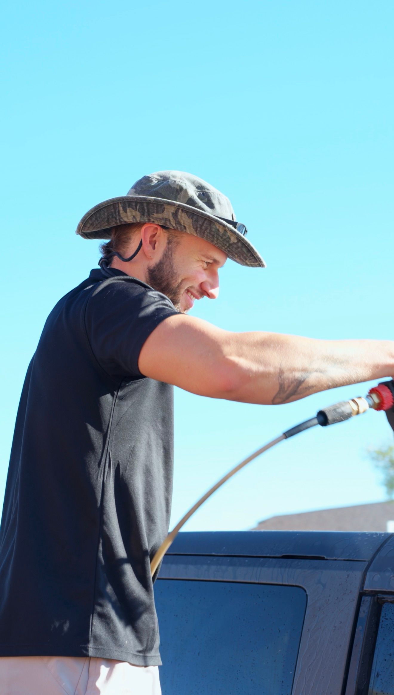 Man in wide-brim hat smiling while using a hose to wash a dark vehicle outdoors on a sunny day.
