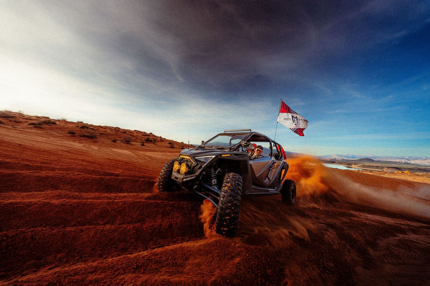UTV racing through red sand dunes kicking up dust with flag flying at golden hour.