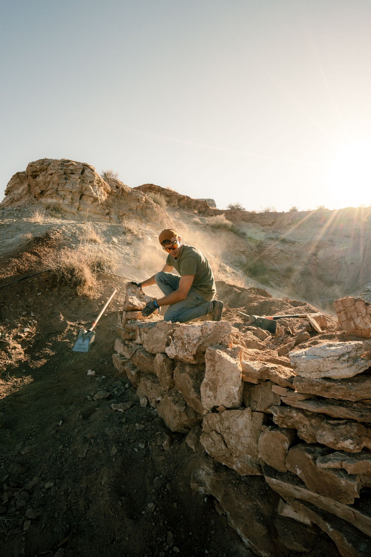 Worker building stone wall on rocky hillside with sunburst and dusty mountain landscape behind him.
