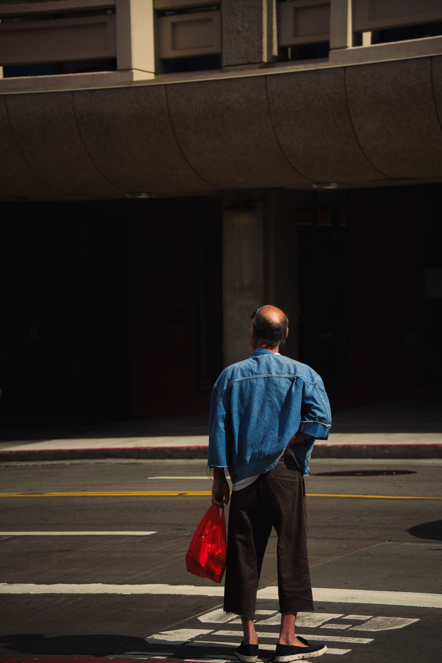 Bald man in blue denim jacket holding a red bag standing barefoot on a sunny urban street corner.