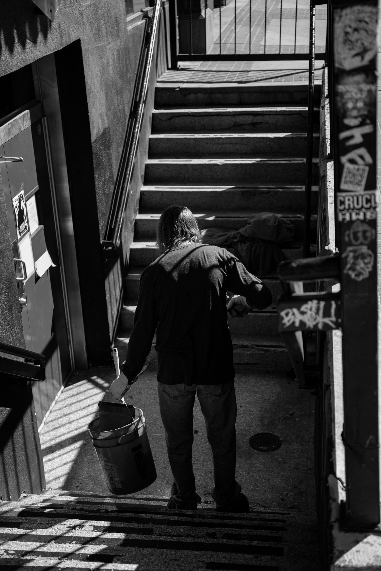 A person carries a bucket toward a stairwell covered in graffiti stickers in black and white urban photography.