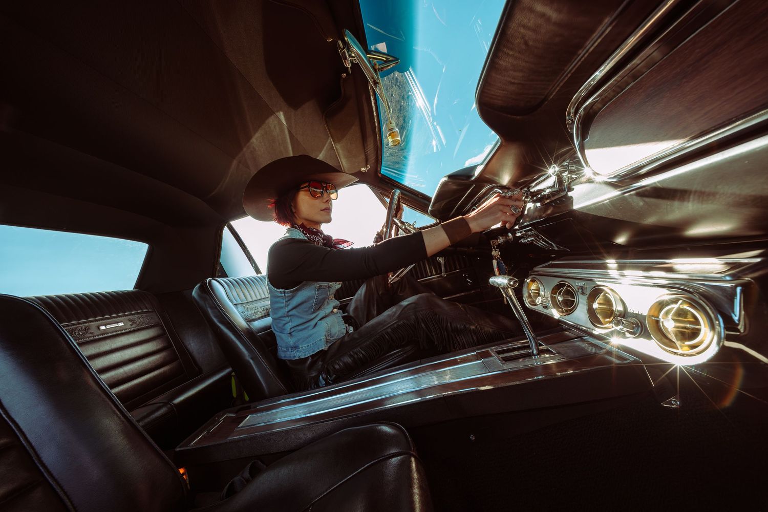 Woman sitting in driver seat of classic vintage car, reaching toward dashboard controls in stylish interior.