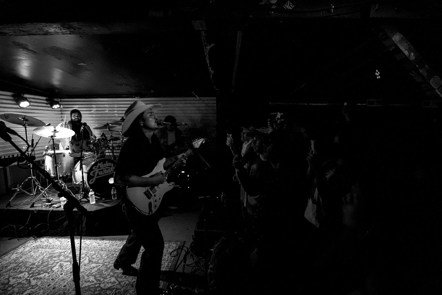 Black and white wide shot of a band performing on stage with drummer and guitarist in a small venue.