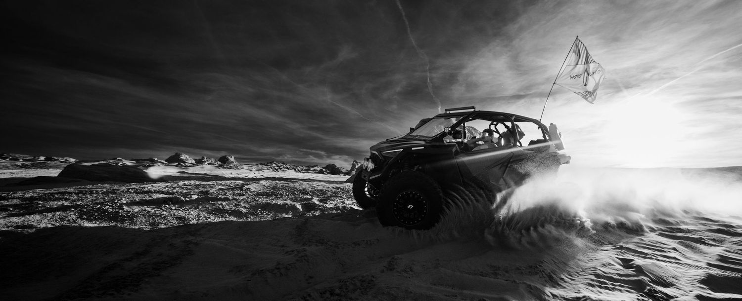 Black and white action shot of an off-road UTV racing through desert sand with dramatic sky.