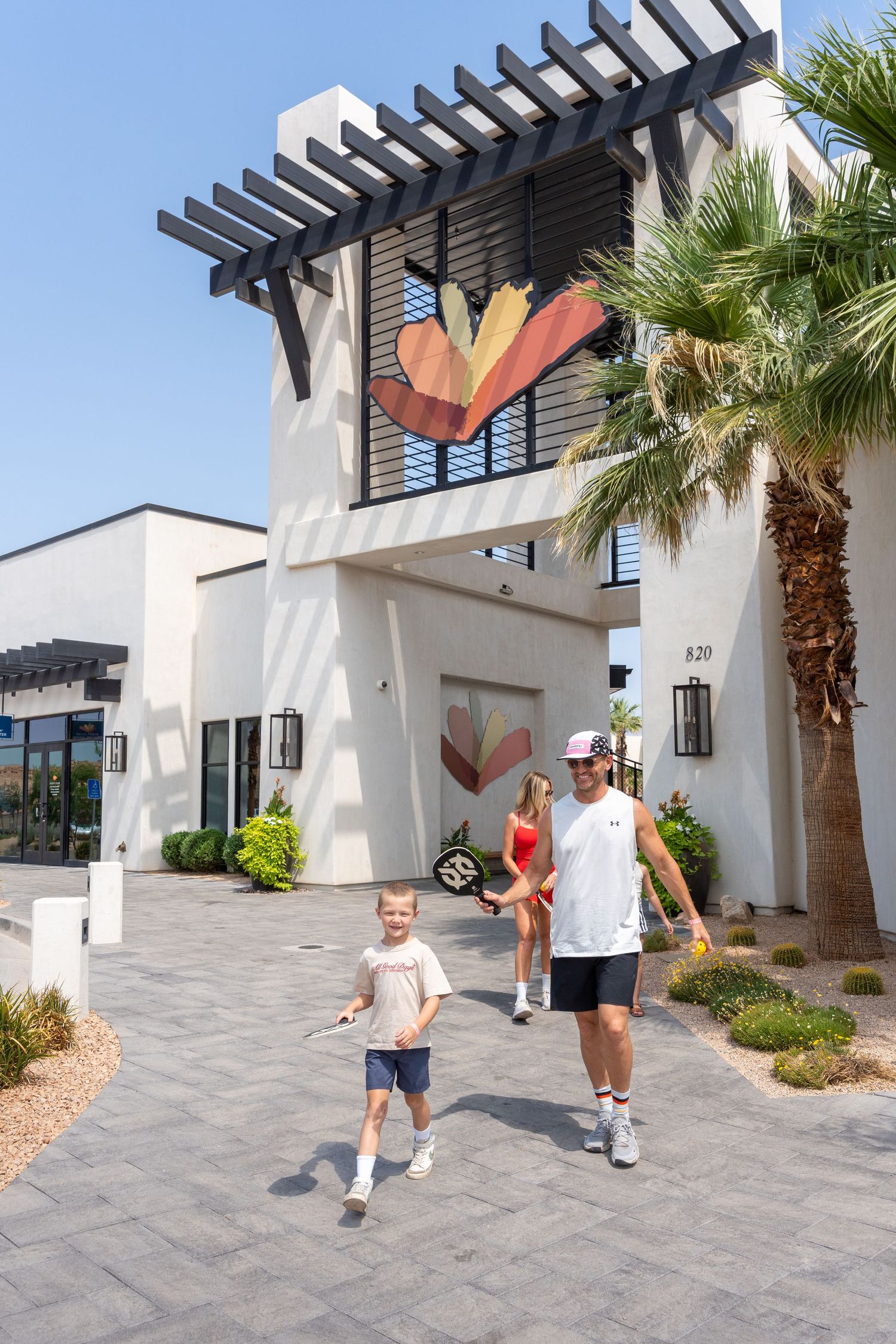 Family walking toward a modern resort entrance with a decorative heart mural and pergola in a sunny desert setting.