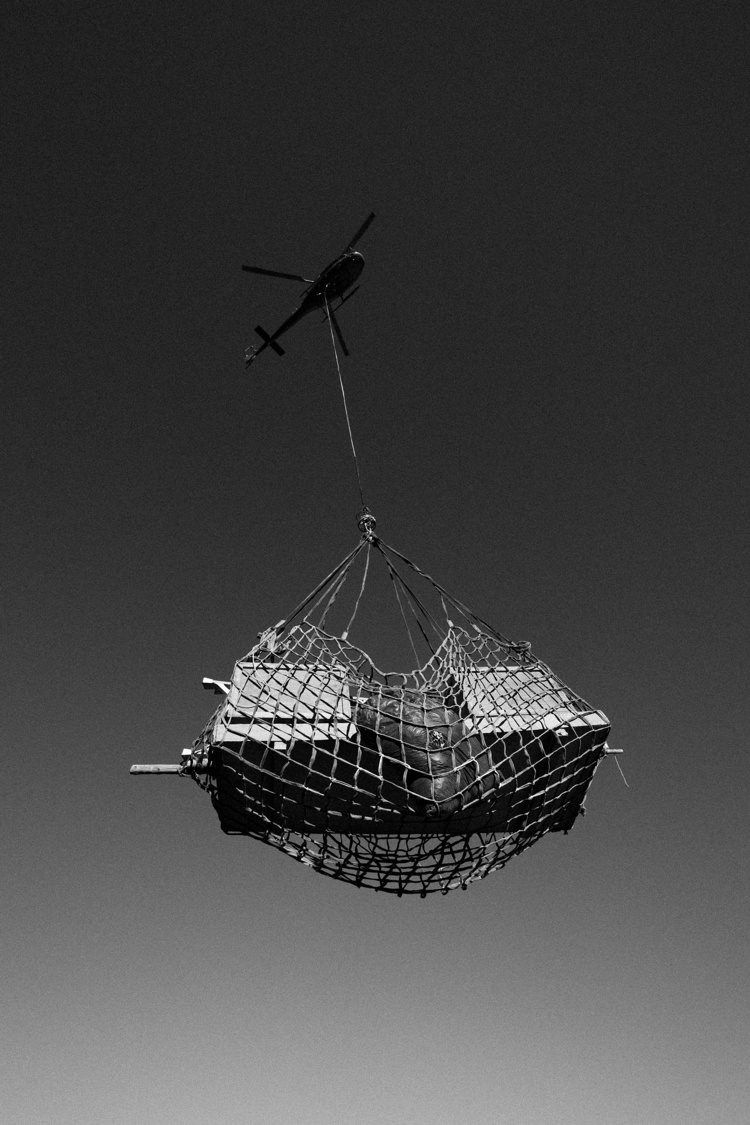 Black and white image of helicopter airlifting cargo in a net against a clear sky.