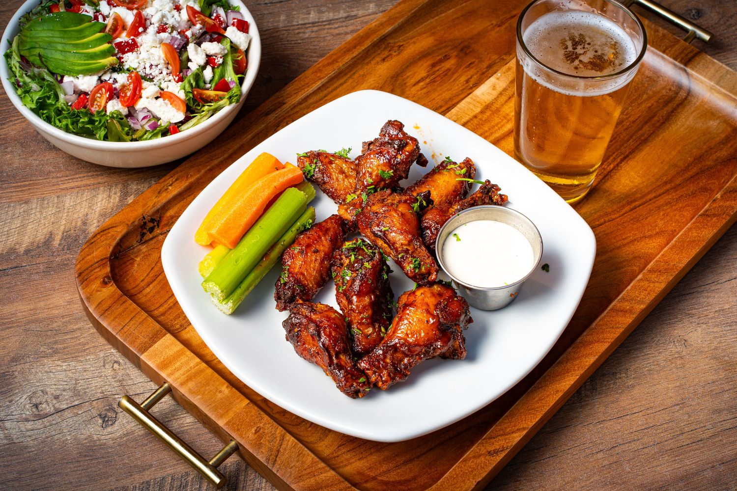 Plate of glazed chicken wings with carrots, celery, and ranch dip served alongside a fresh salad and beer.