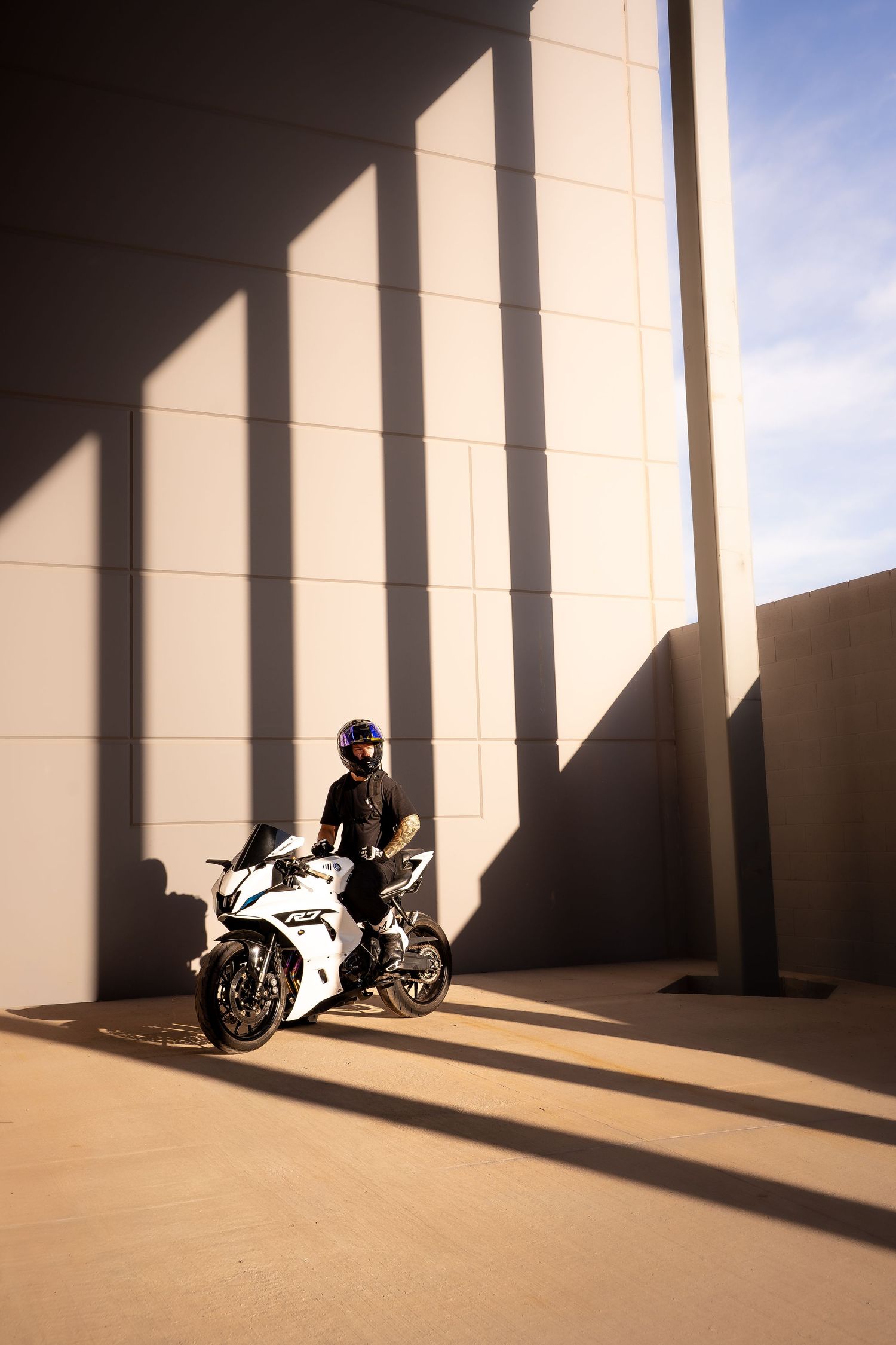 Rider on white Yamaha R1 motorcycle poses in sunlit concrete corridor with bold geometric shadows.
