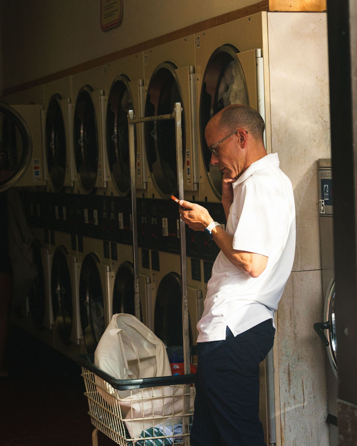A bald man checks his phone while standing beside stacked washing machines in a laundromat.