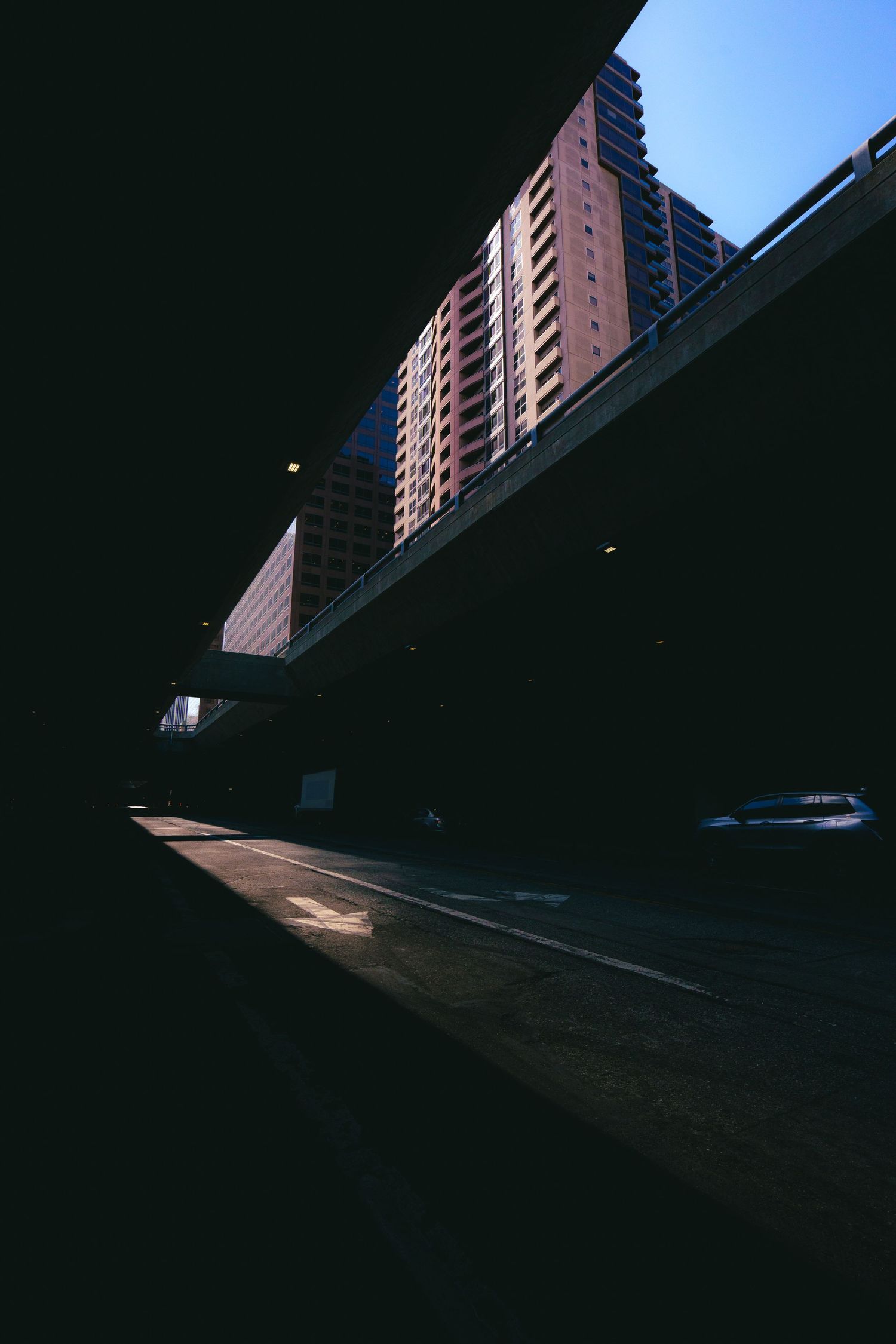 Dark urban street scene under an overpass with a tall residential building illuminated by sunlight.