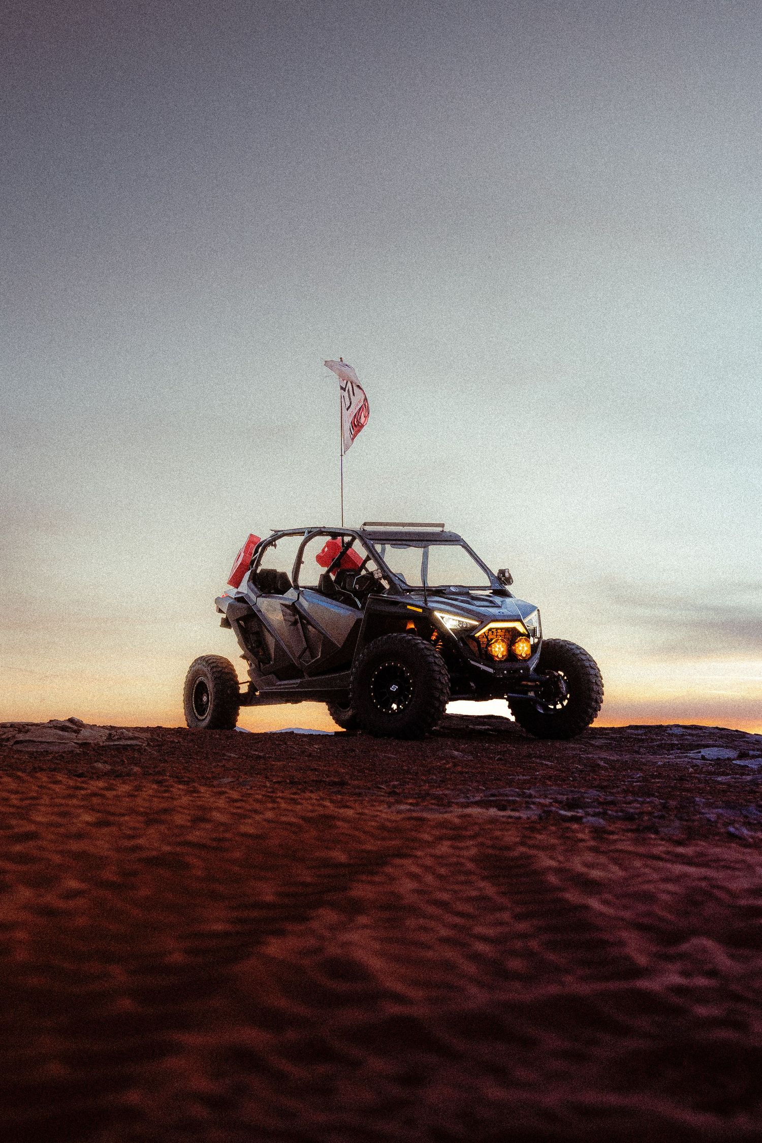 A Can-Am UTV side-by-side with a flag parked on desert sand dunes at sunset.