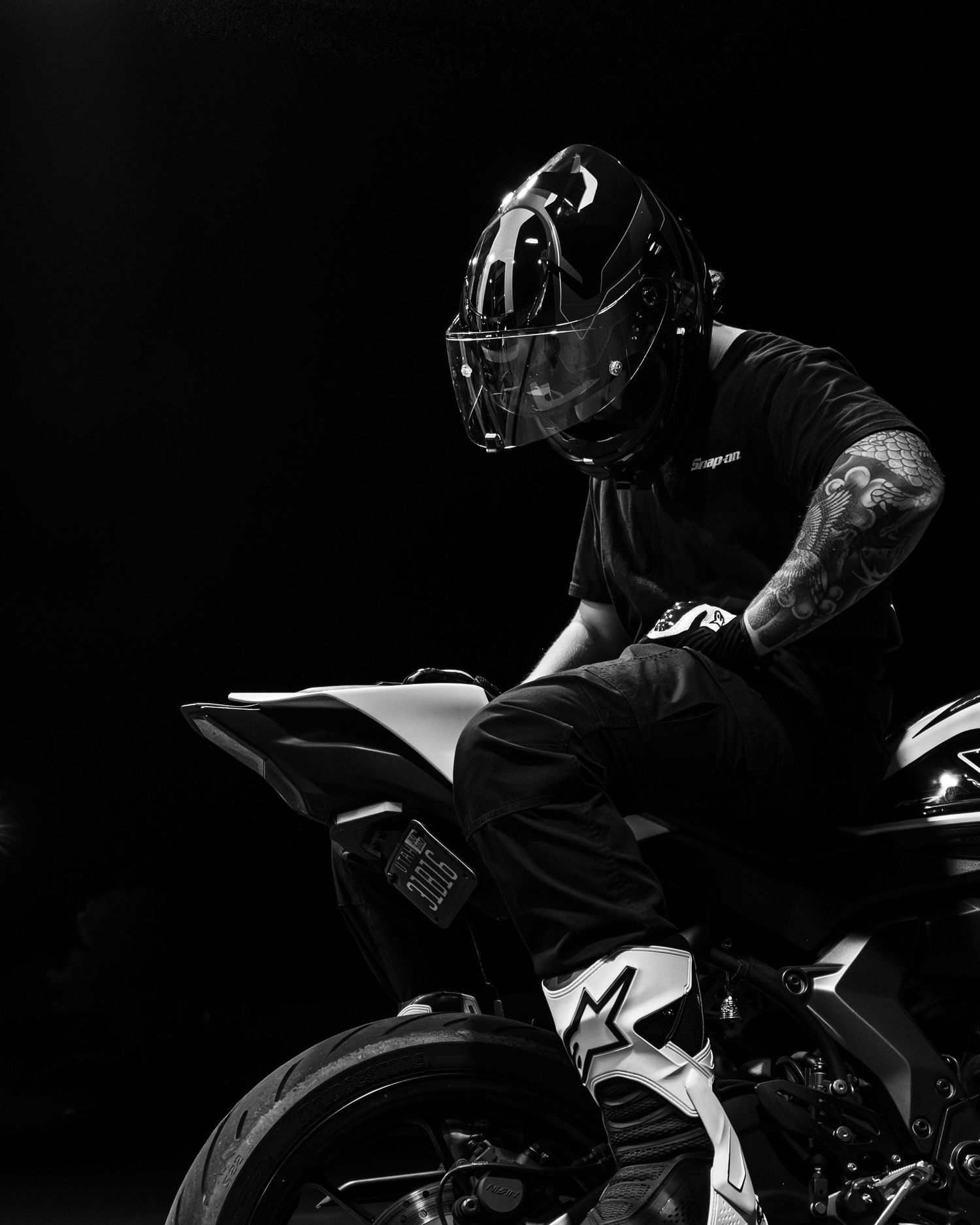 Black and white photo of tattooed rider in full-face helmet crouched over sport motorcycle.