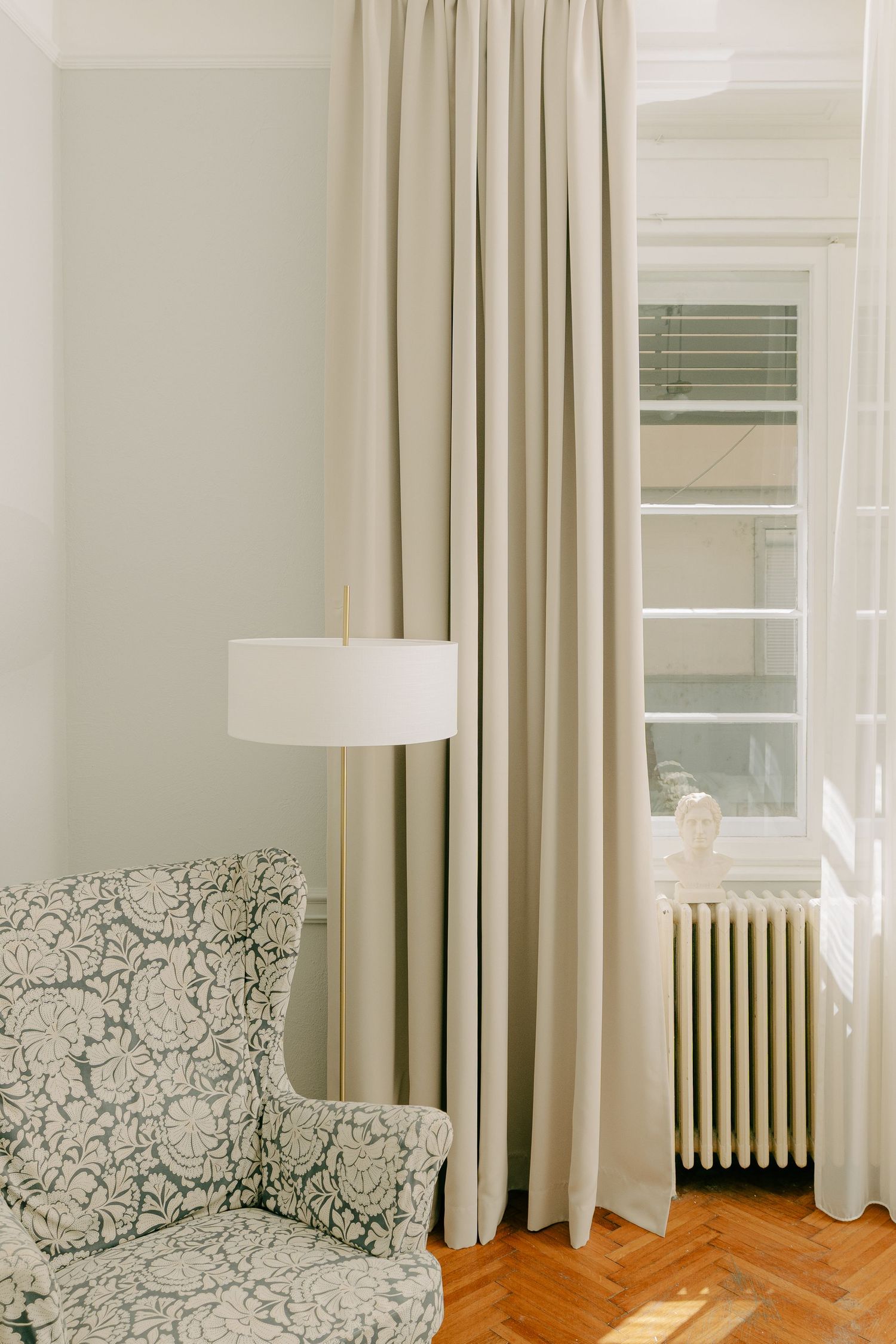 Elegant living room corner with floral armchair, floor lamp, beige curtains, and vintage radiator by a sunlit window.