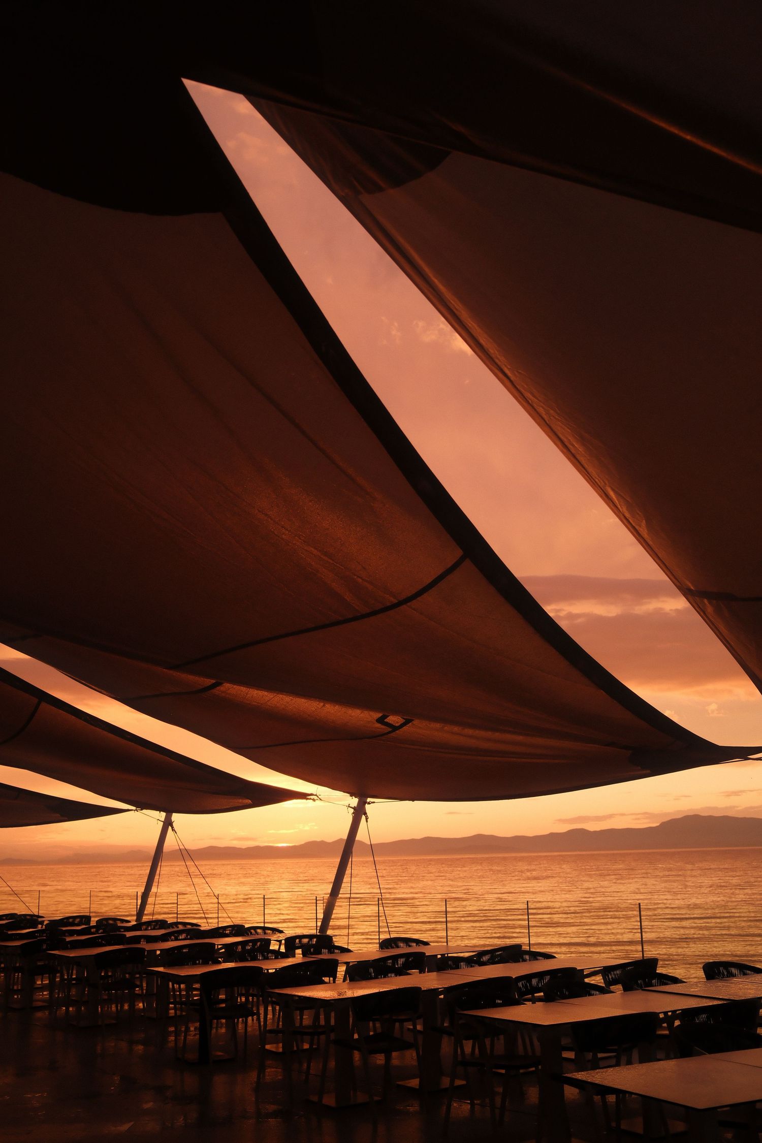 Outdoor beach restaurant with sail shades at sunset, tables silhouetted against golden orange sky over calm ocean waters.