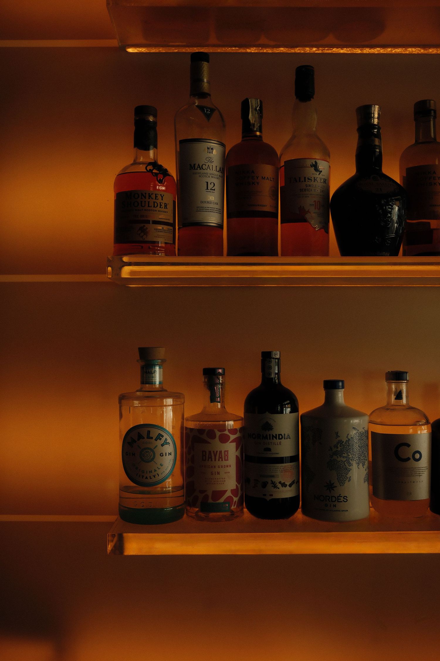 Dimly lit wooden shelves displaying various liquor bottles in warm ambient lighting creating a cozy bar atmosphere.