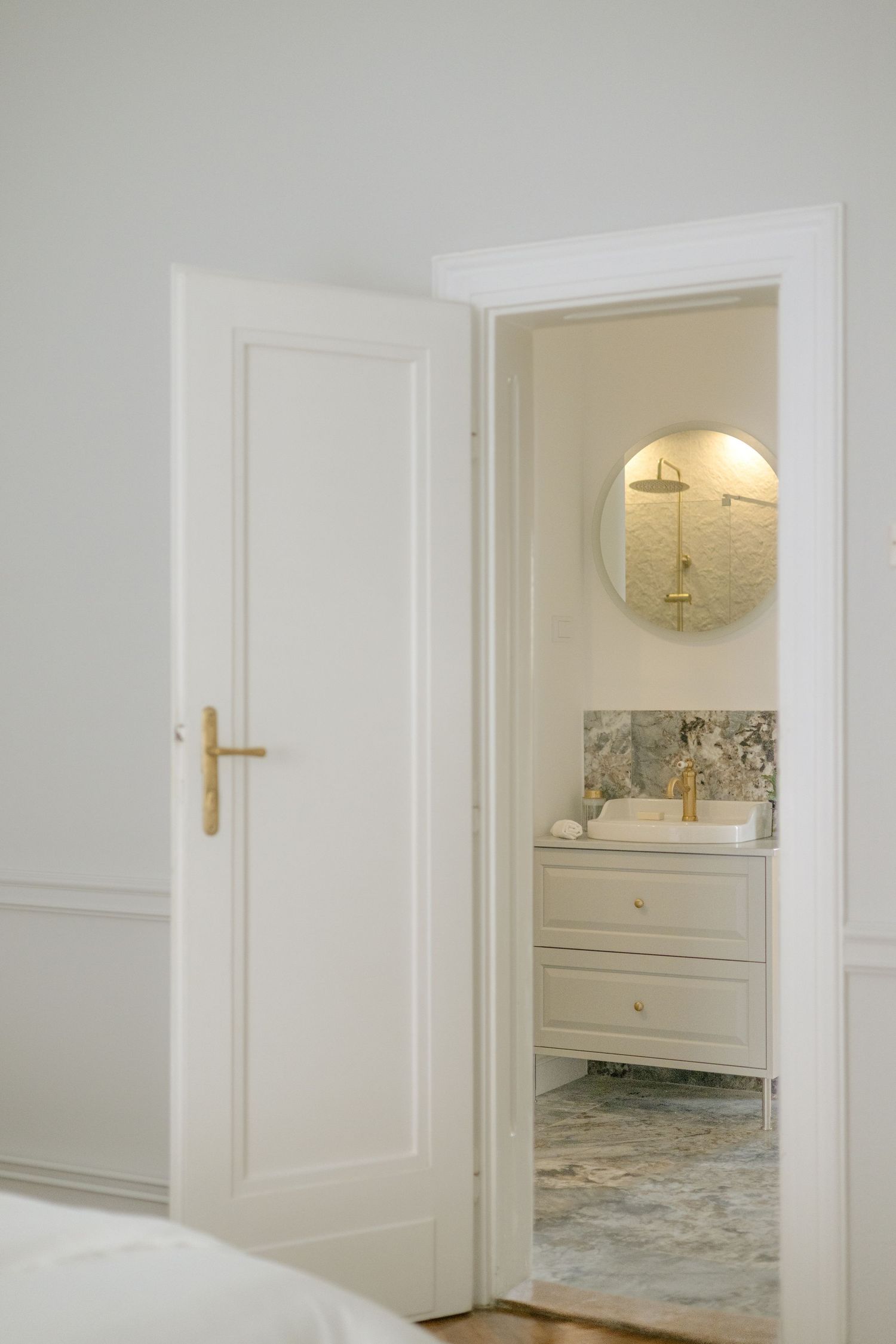 Elegant white bedroom doorway opening to a luxurious en-suite bathroom with gold fixtures and round mirror.