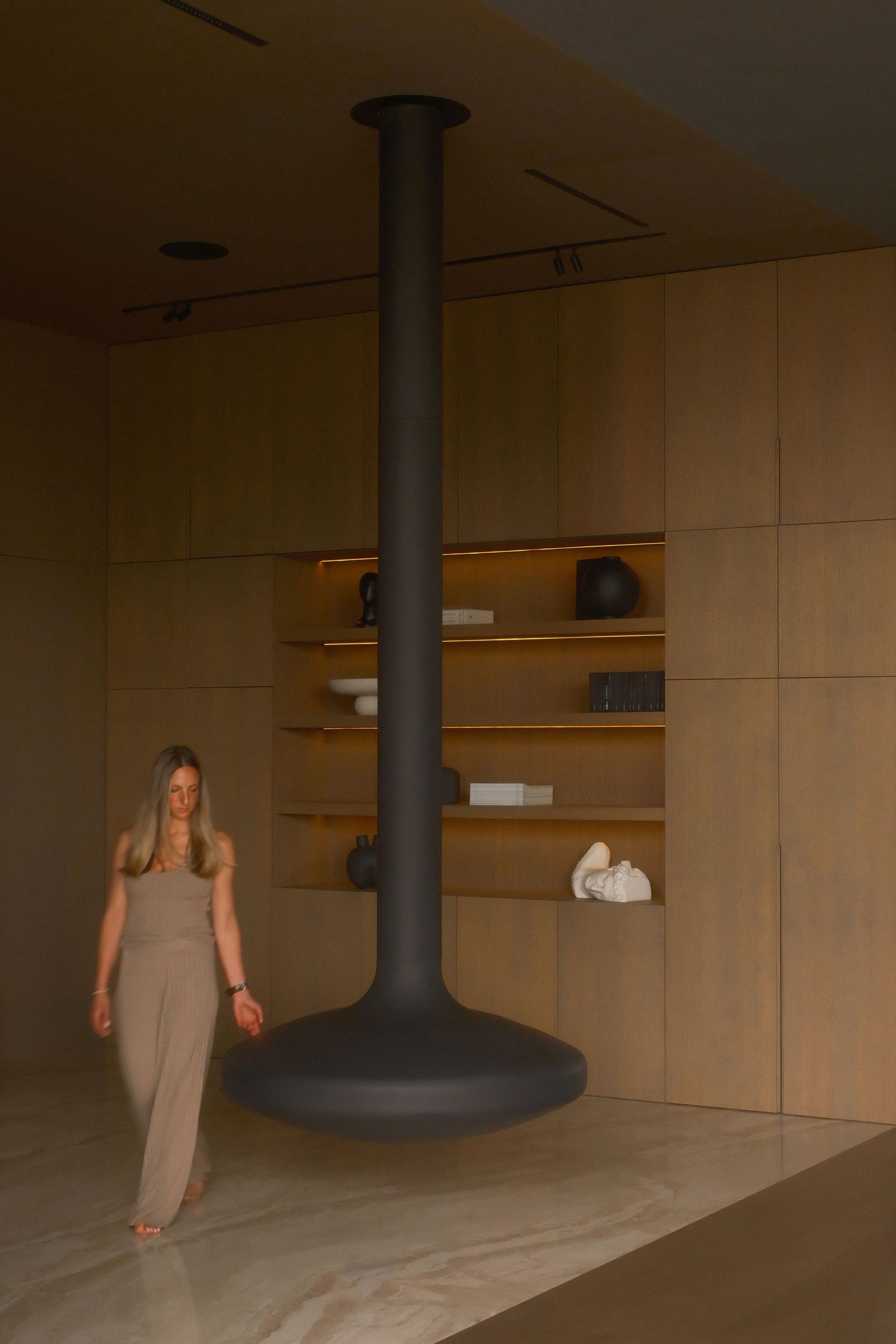 Woman in beige dress stands beside a modern black pendant fireplace in a minimalist wood-paneled showroom.